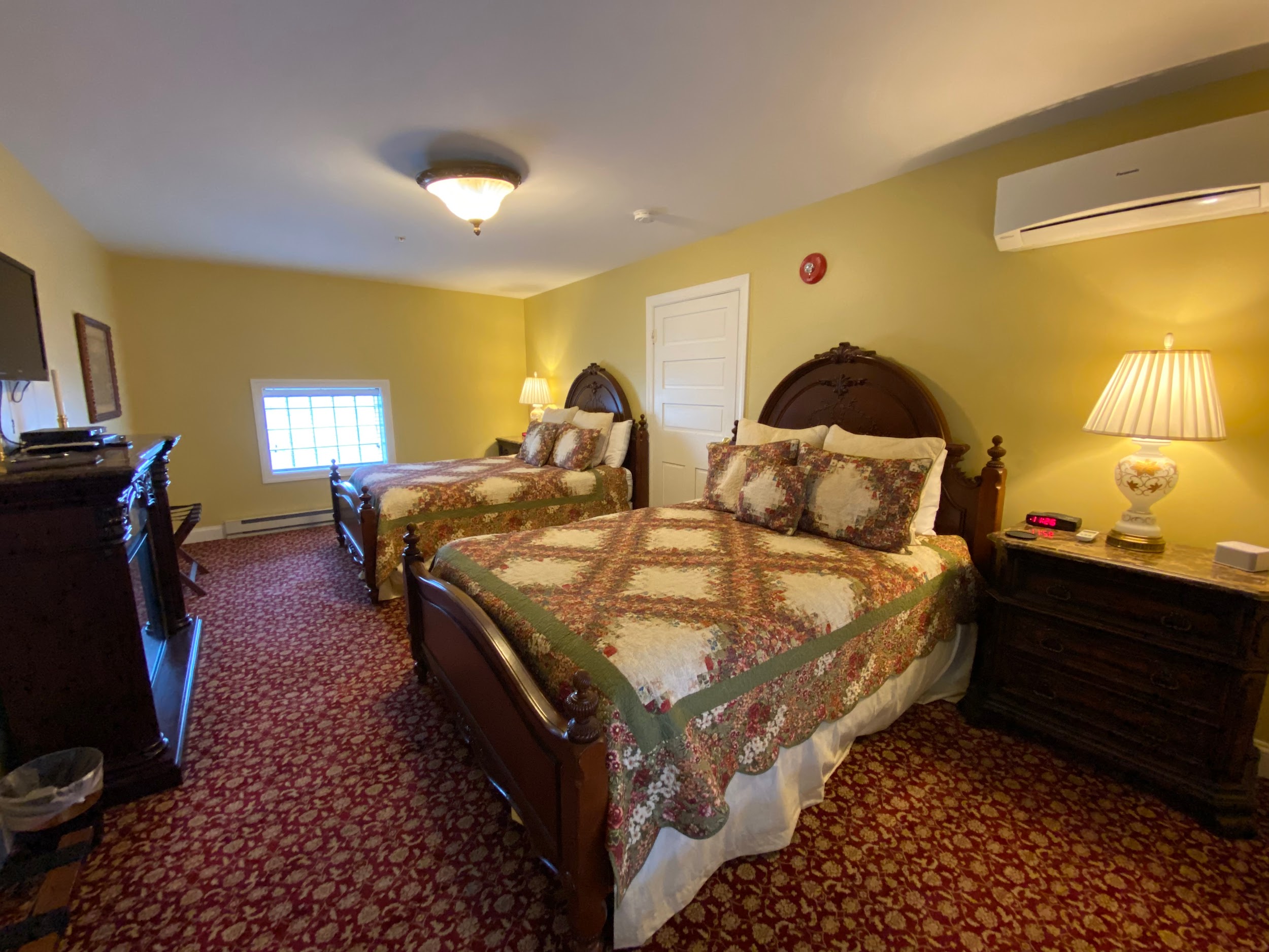 The Swope Suite features a carpeted bedroom, 2 queen-size beds with wooden headboards, 2 night stands with lamps, an electric fireplace, and a split HVAC unit.