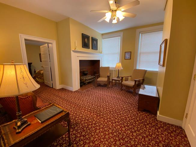 A sitting room with yellow walls, a red patterned carpet, a fireplace with a white mantle, two armchairs facing the fireplace, a small table with a lamp, and two windows.