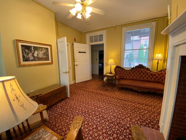 A sitting area with yellow walls, a red patterned carpet, a Victorian sofa, a lamp, a framed picture, and a doorway leading to another room.