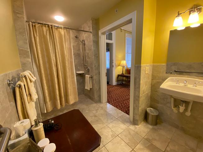 The Swope Suite bathroom is ADA accessible, with a roll in shower with grab bars, tile floor, roll under sink, toilet with grab bars, a table for toiletries.