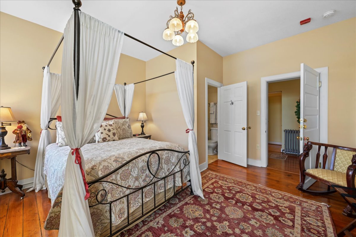 The Northern Spy Chamber features a queen-size metal canopy bed, a dresser, 2 night stands with lamps, 2 Victorian chairs, an electric fireplace, original wooden floors, and an area.