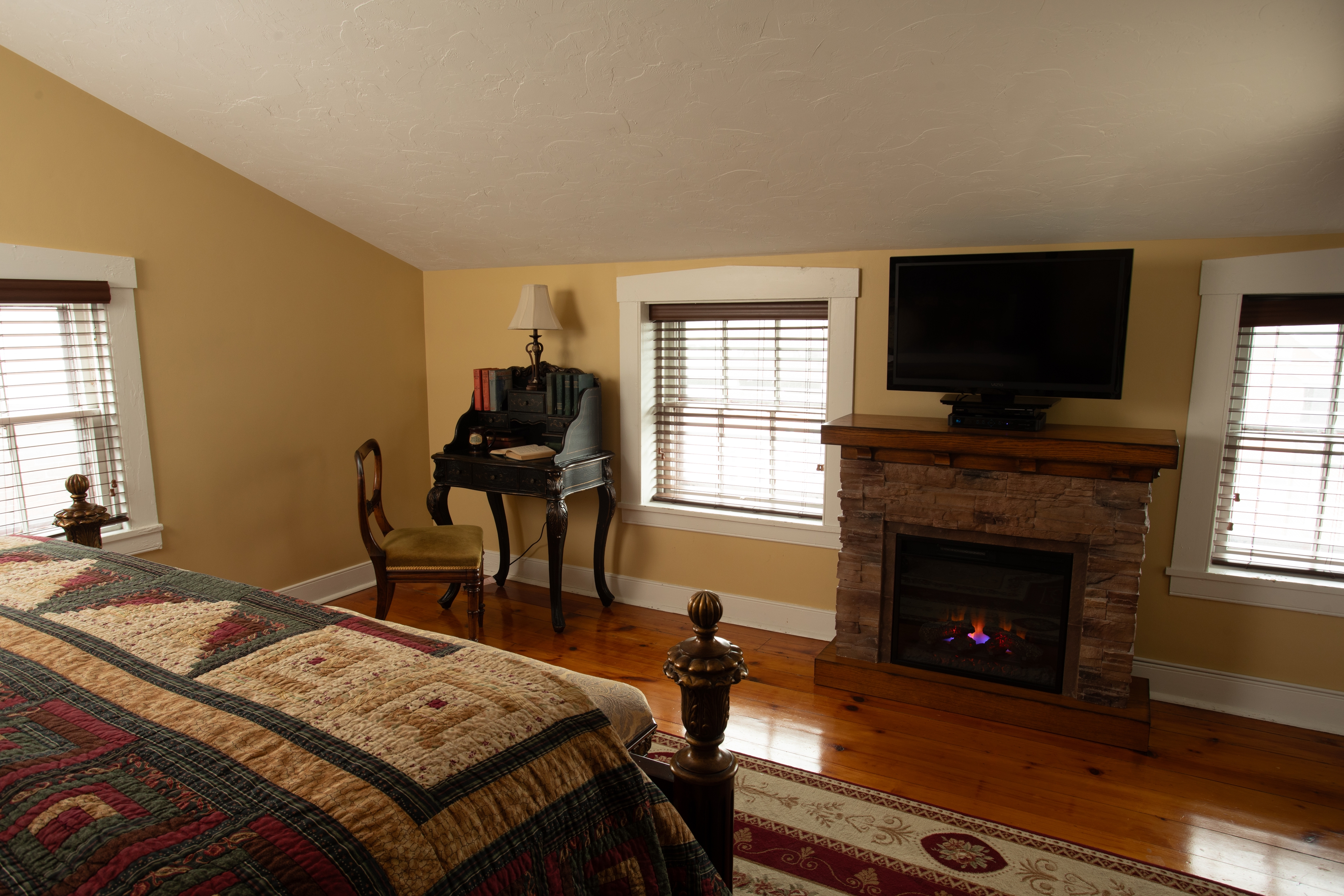 Cozy bedroom featuring a quilted bed, a fireplace, a TV, and a writing desk.