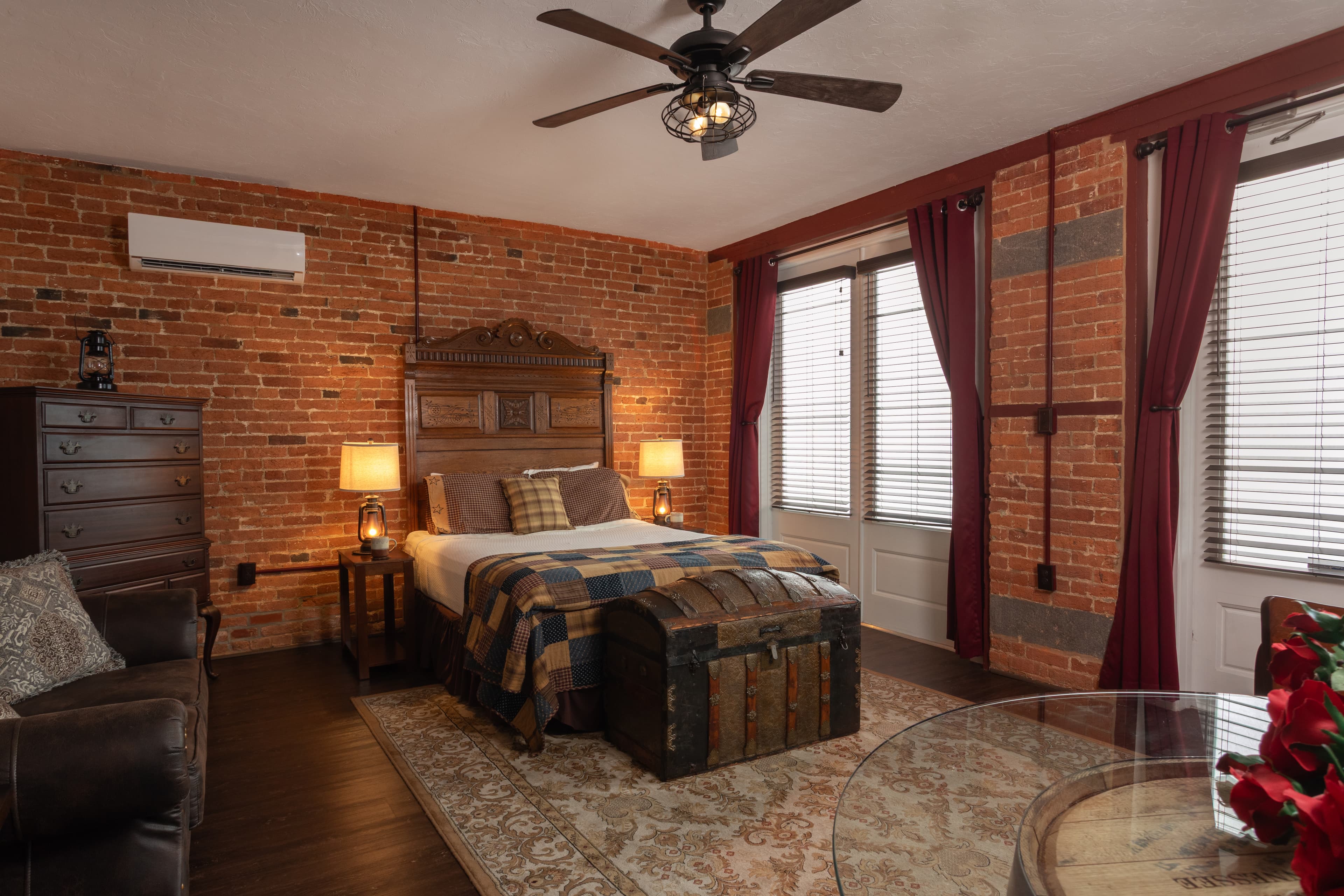The Carriage House bedroom featuring exposed brick walls, a bed with plaid bedding, and large windows with red curtains.