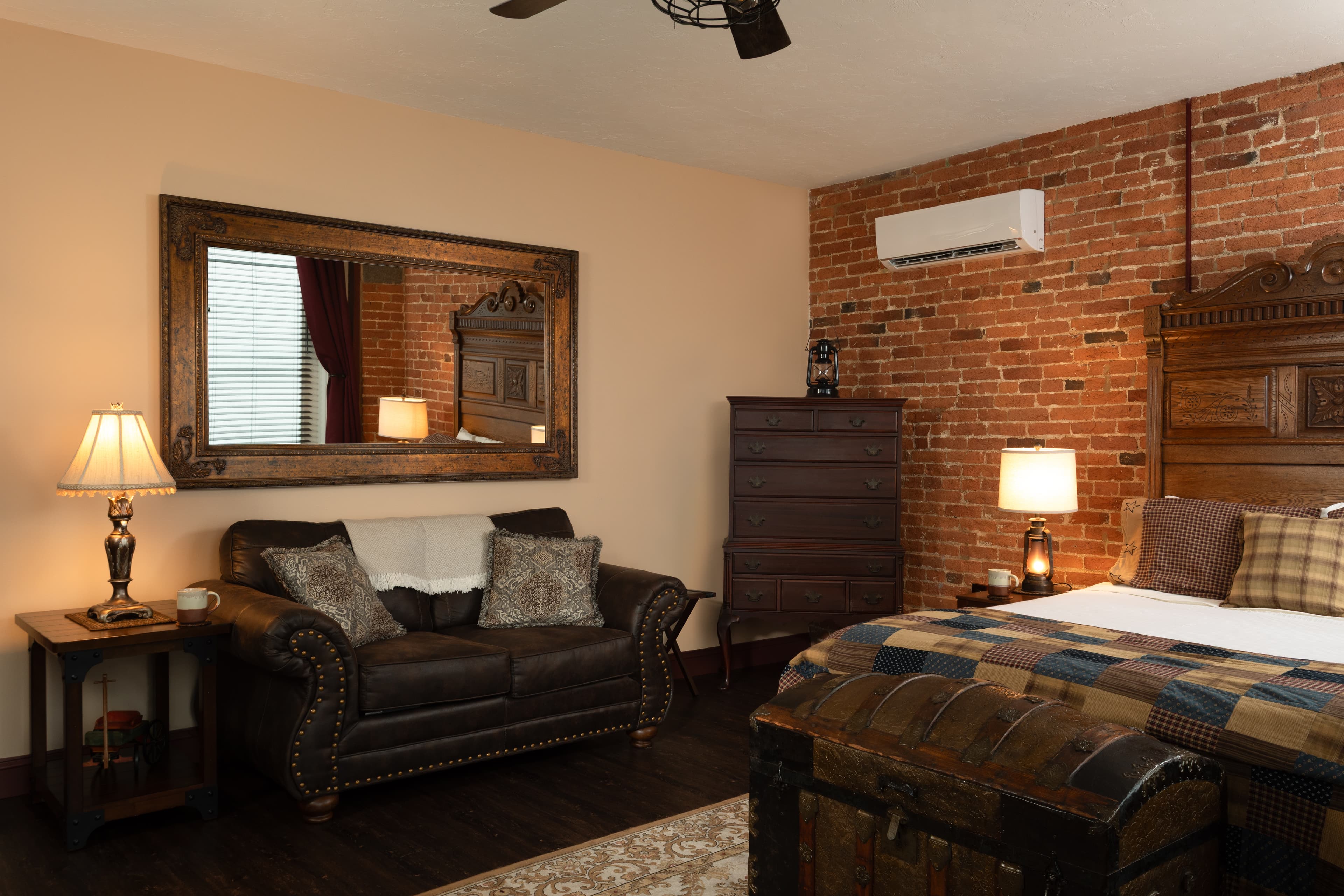 The Carriage House bedroom featuring a leather couch, wooden dresser, and a large mirror against a brick wall.