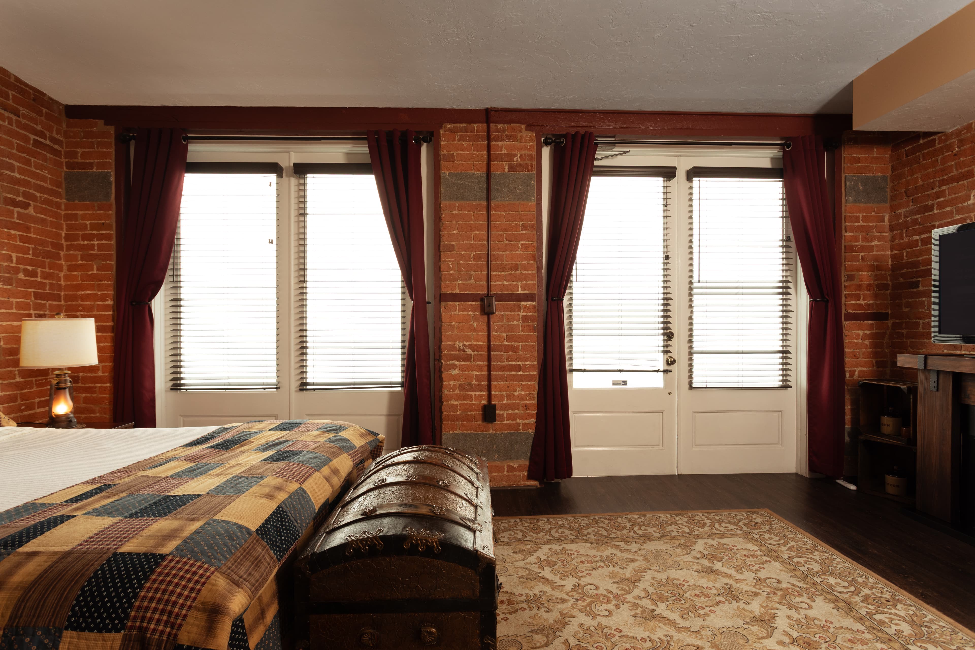 The Carriage House bedroom featuring brick walls, dark curtains, a patterned blanket, and large windows.
