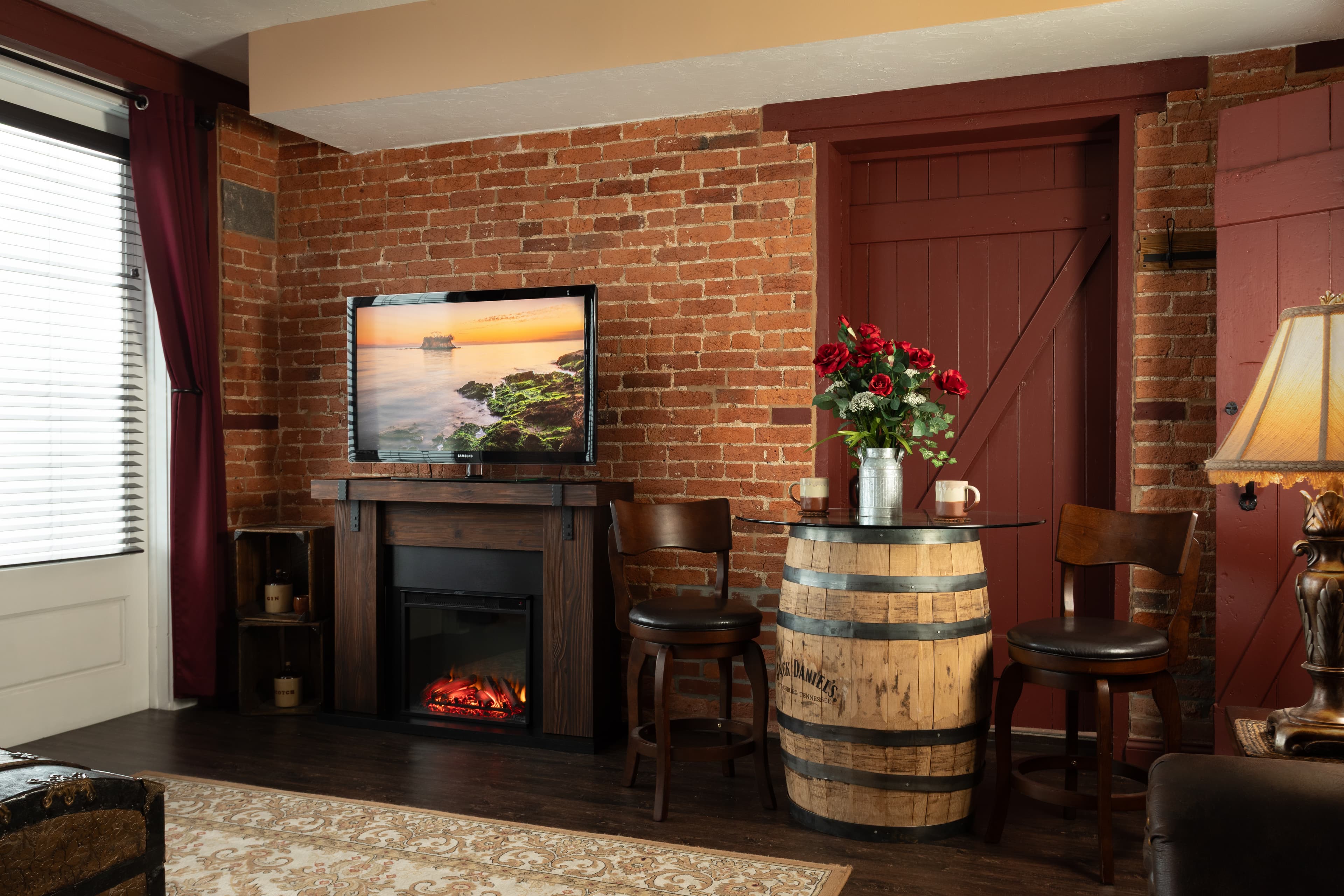 The Carriage House living room with a brick wall, a flat-screen TV, an electric fireplace, and a barrel table with flowers.