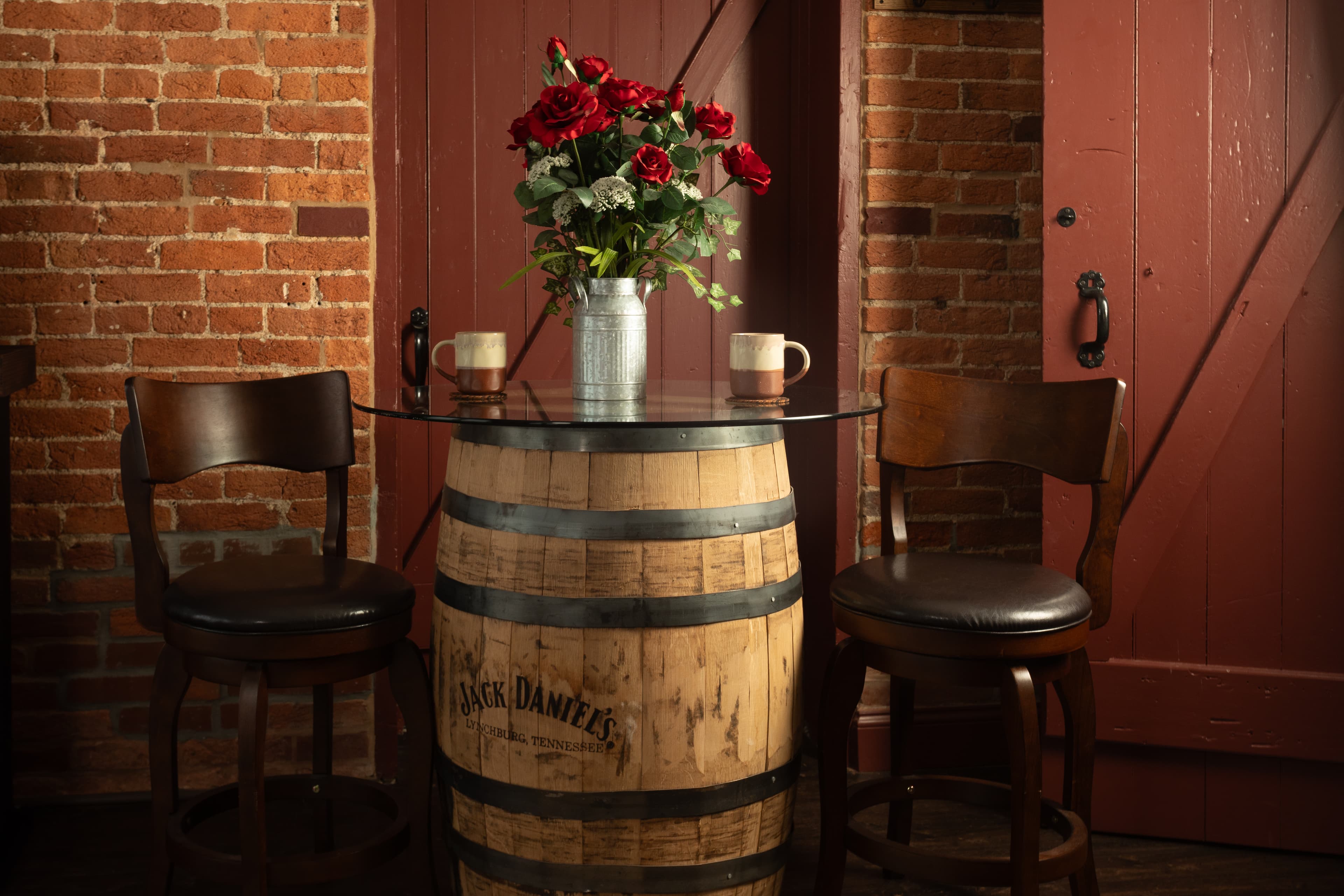 A rustic table made from a whiskey barrel topped with a glass surface, adorned with a vase of red roses and two mugs, set against a brick wall in the Carriage House.