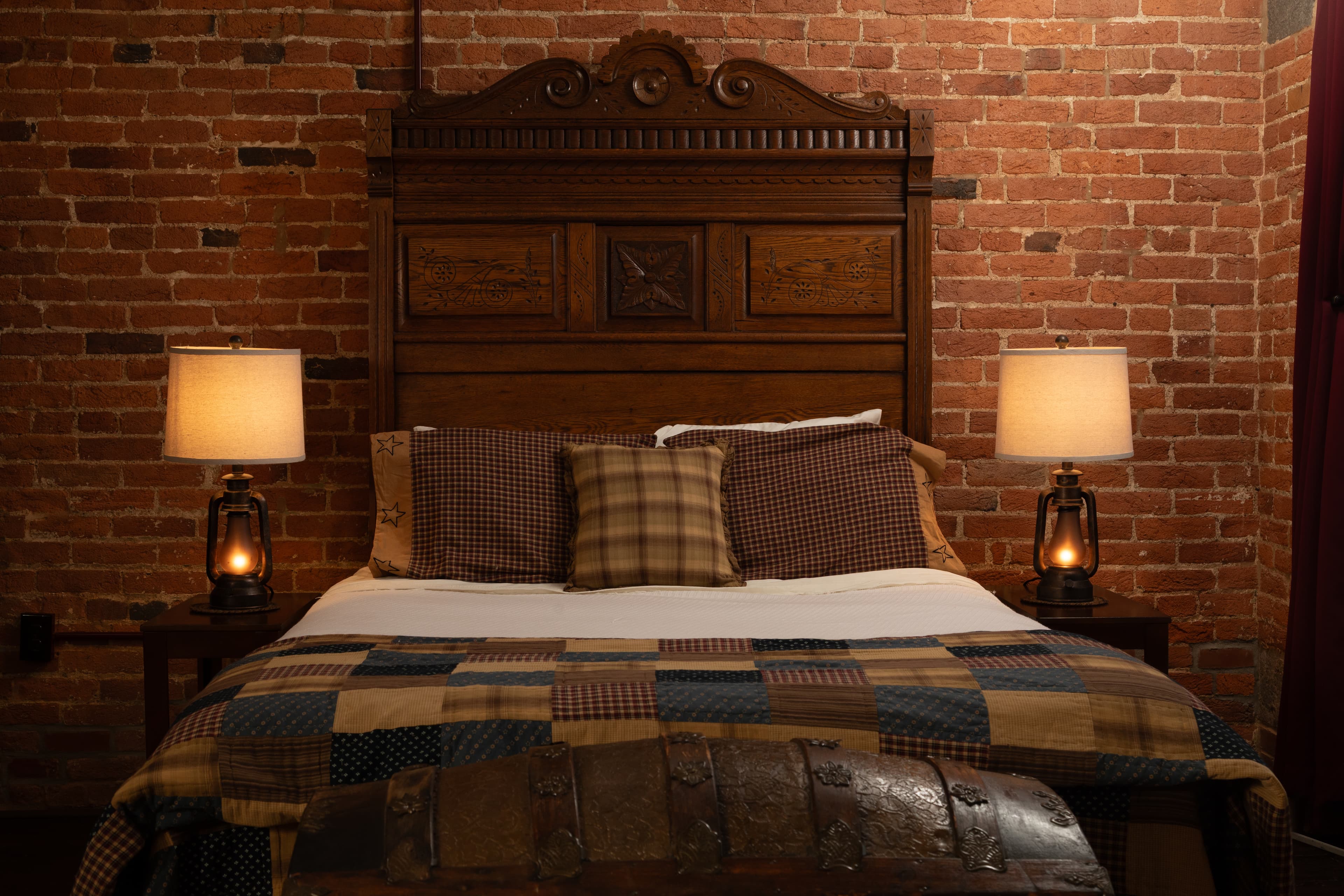 The Carriage House bedroom featuring a wooden bed, plaid bedding, and two bedside lamps against a brick wall.