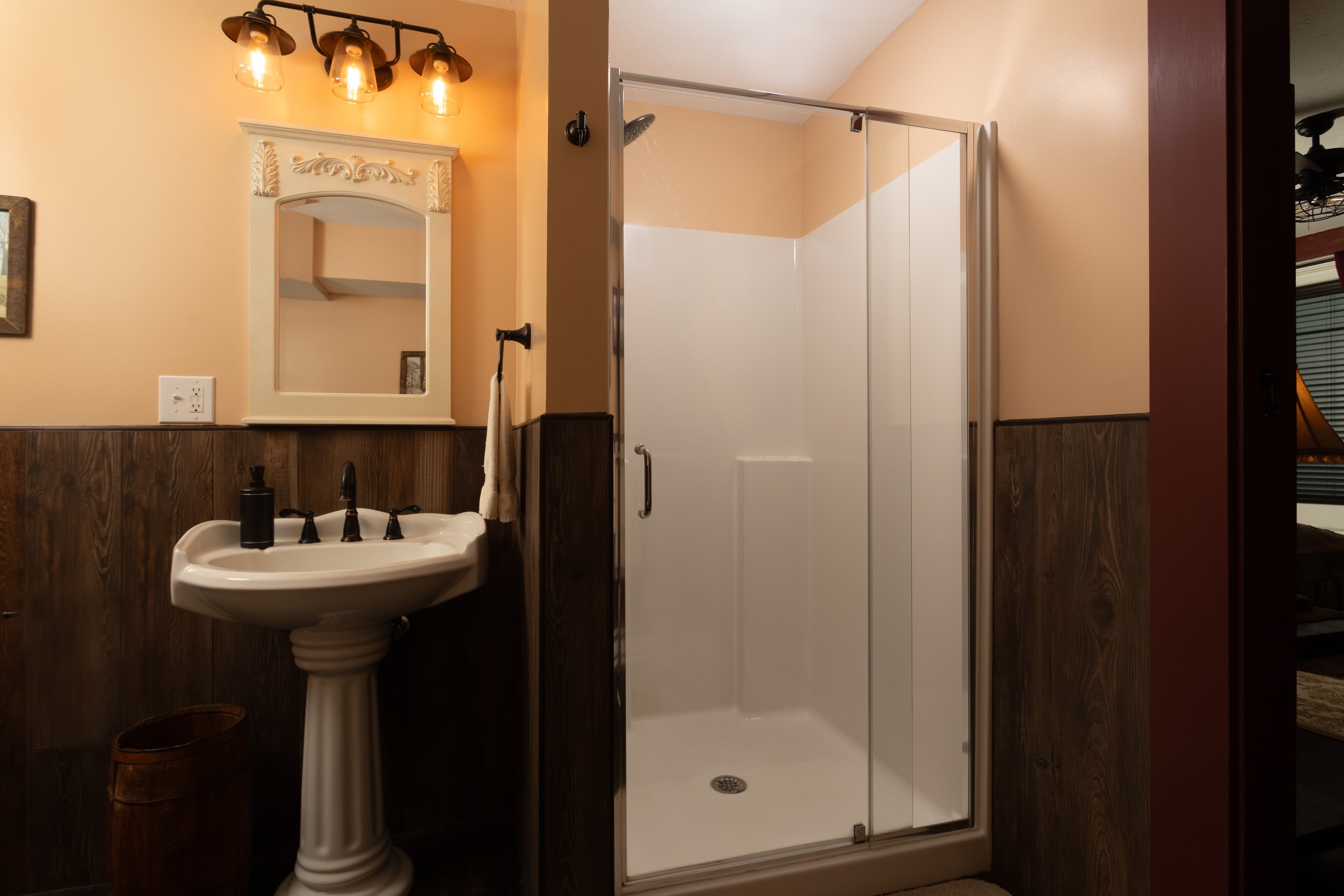 A modern bathroom in the Carriage House featuring a glass-enclosed shower, pedestal sink, and warm lighting.