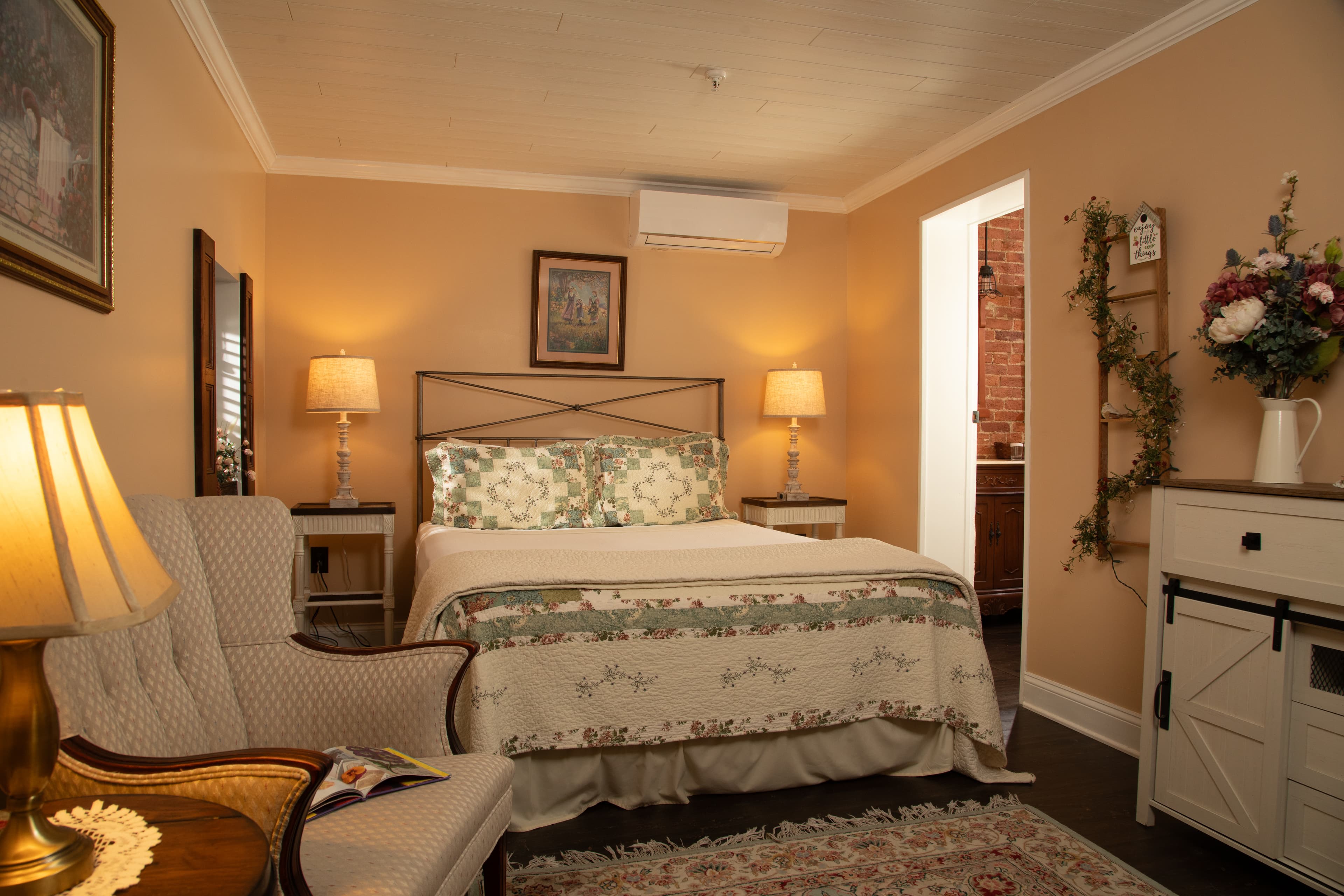 Cozy bedroom with a double bed, soft lighting, and rustic decor, featuring an armchair and a doorway leading to another room.