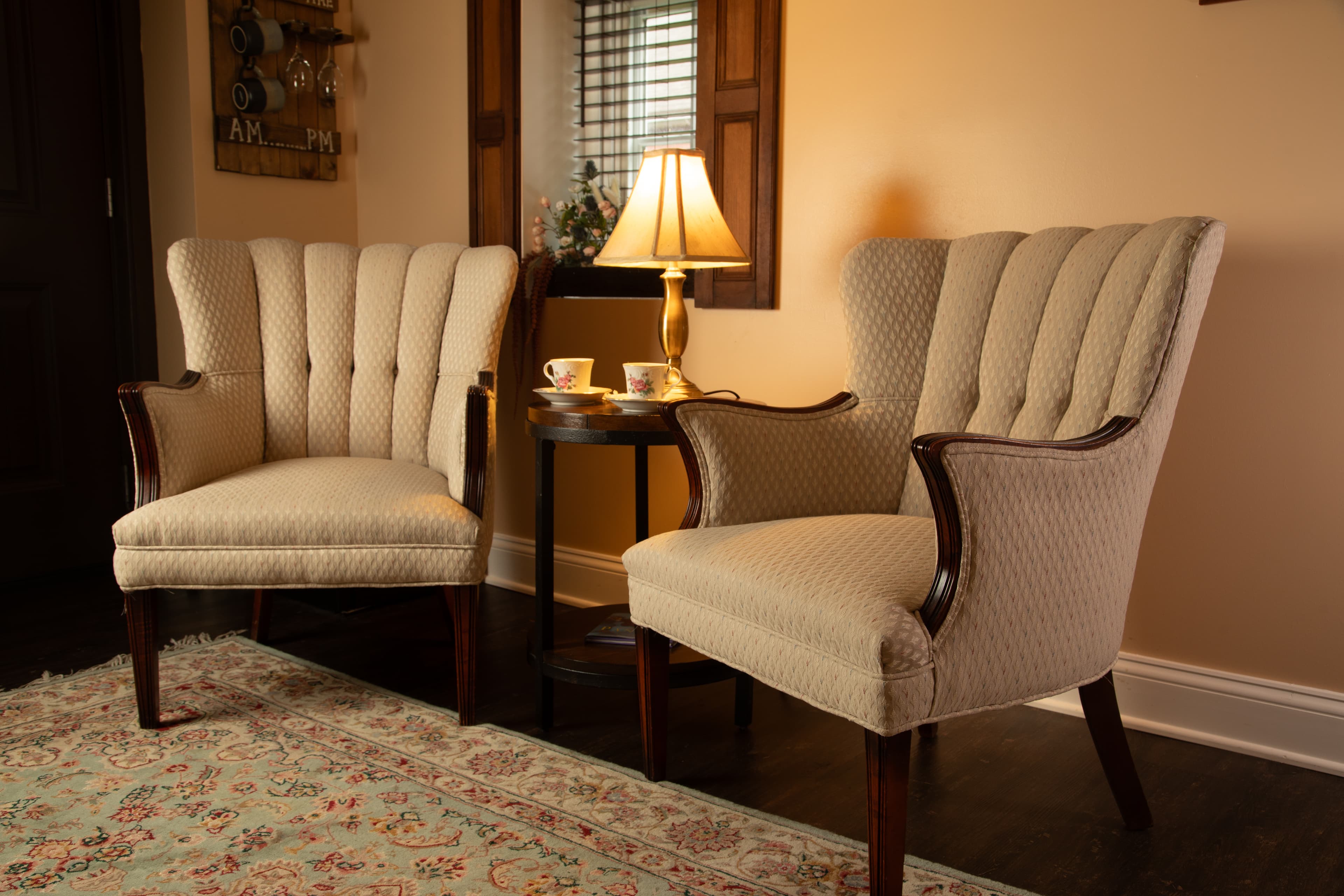 Two beige upholstered armchairs flanking a small table with a lamp and tea cups, set in a cozy living room.