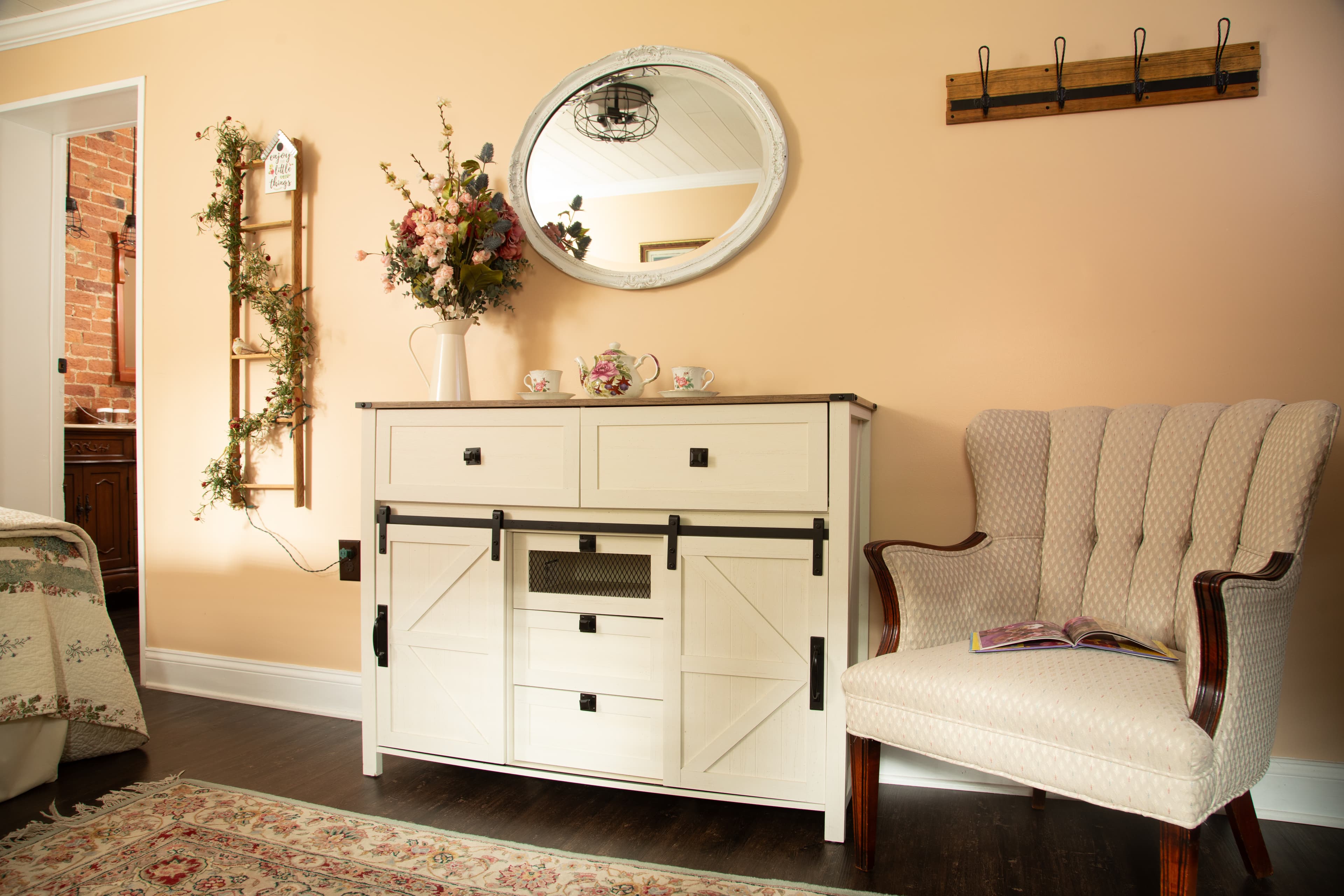 A cozy living space featuring a white sideboard, a vintage armchair, a round mirror, and decorative flowers against a pastel wall.