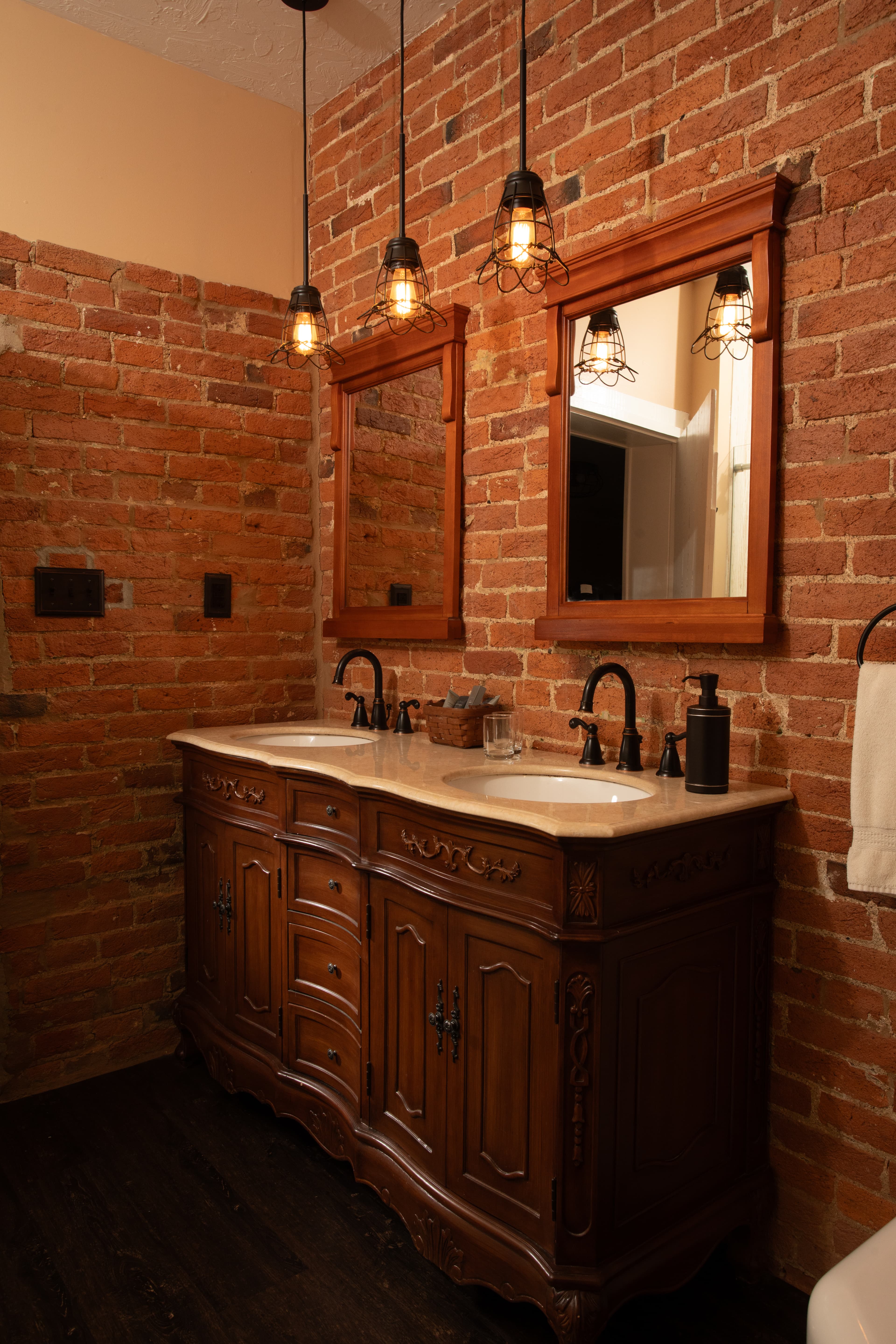 A rustic bathroom featuring a wooden double vanity, brick walls, and pendant lighting.