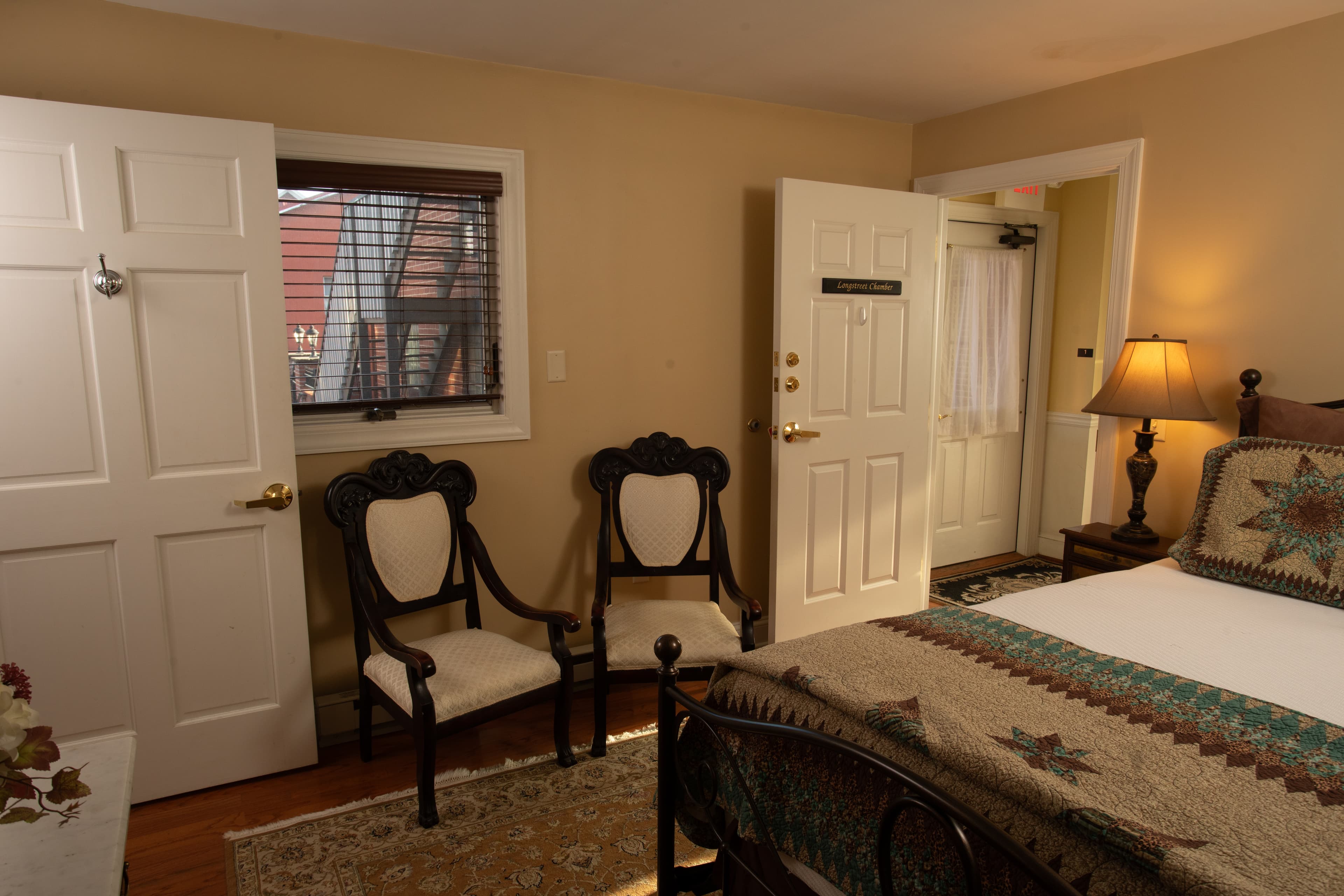 Cozy bedroom featuring a bed with a quilt, two chairs, a lamp, and doors leading to the hallway and outside.