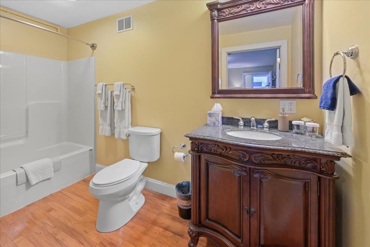 The Longstreet Chamber bathroom features wooden floors, an ornate wooden sink cabinet with marble top, toilet, and a tub / shower
