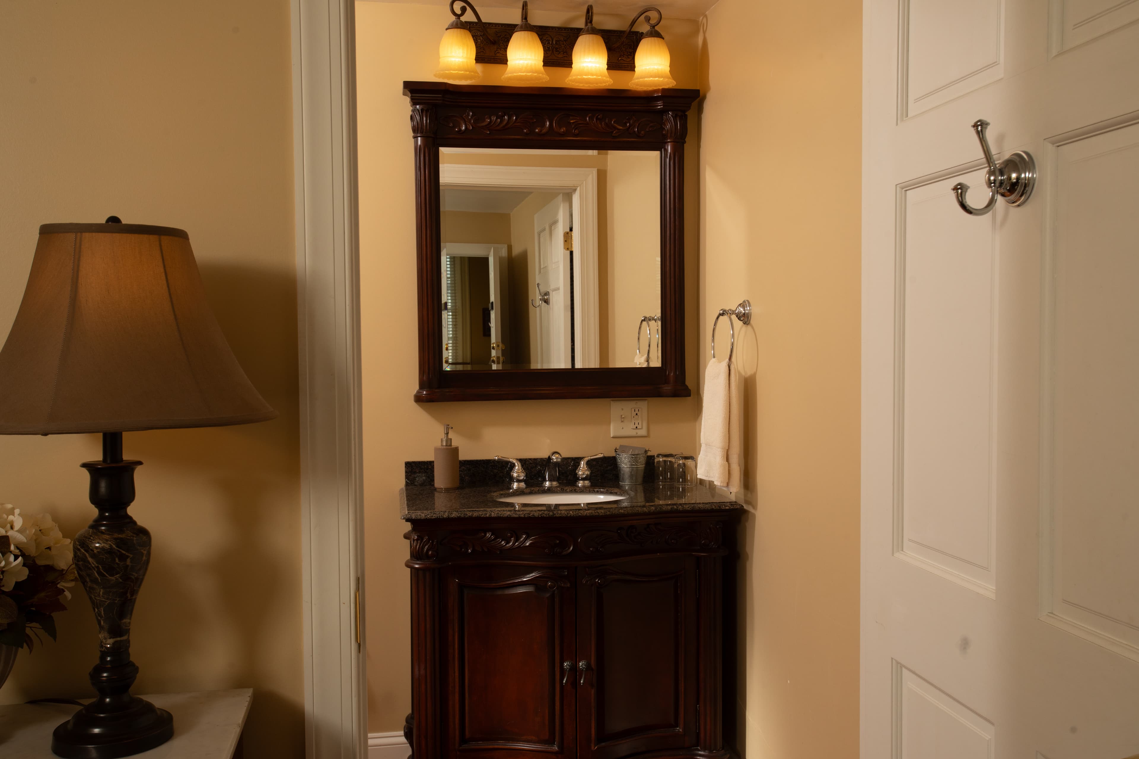 A bathroom featuring a dark wood vanity, a mirror with lights above, a lamp, and decorative elements.