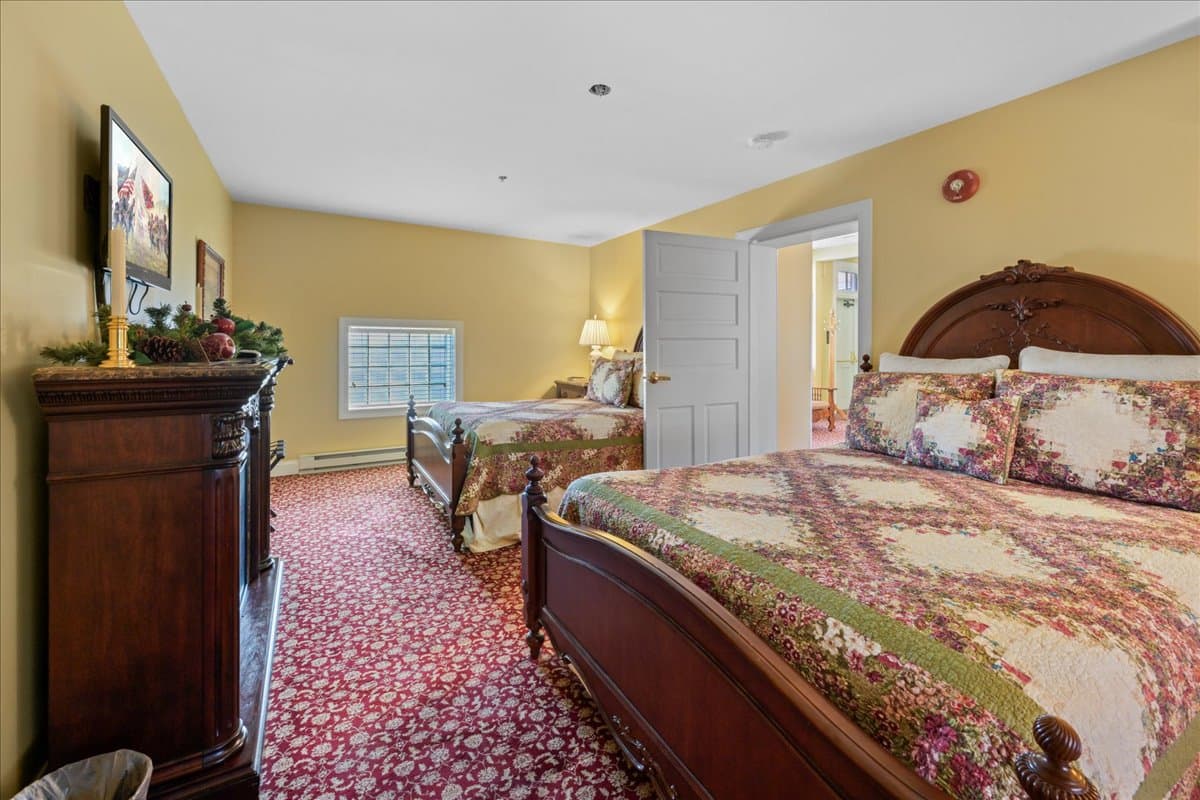 A sitting room with yellow walls, a red patterned carpet, a fireplace with a white mantle, two armchairs facing the fireplace, a small table with a lamp, and two windows.