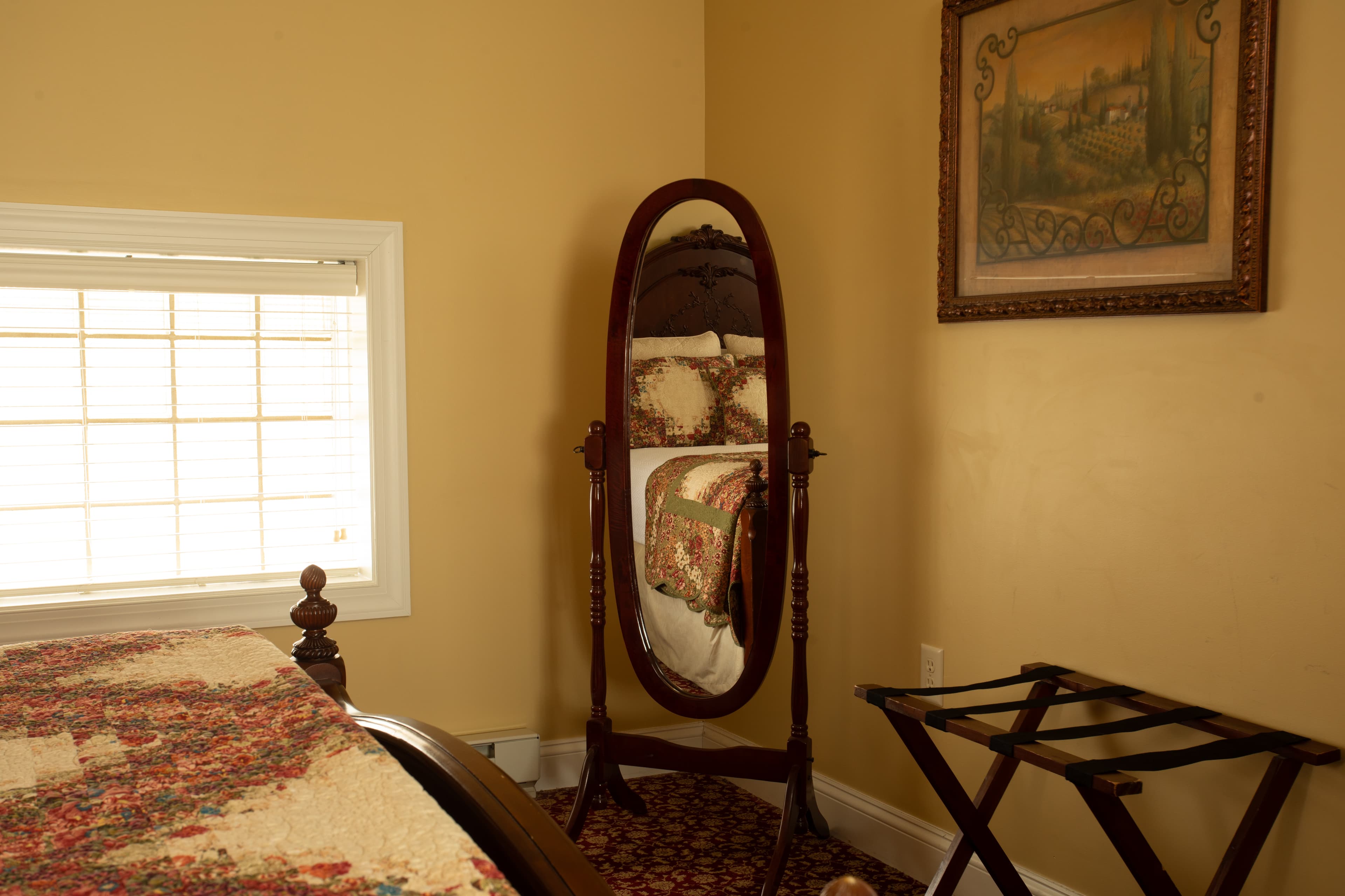A cozy bedroom corner featuring a wooden mirror, a quilted bed, a small folding table, and a painting on the wall.