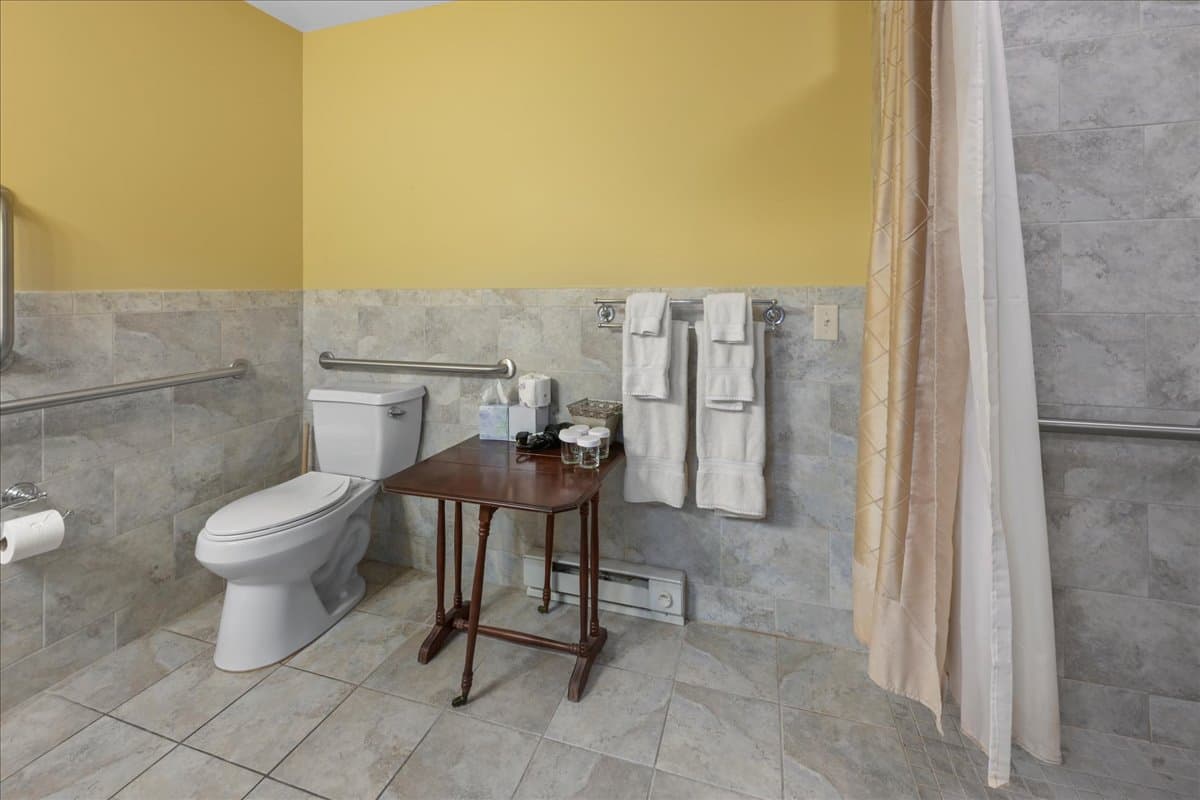 The Swope Suite bathroom is ADA accessible, with a roll in shower with grab bars, tile floor, roll under sink, toilet with grab bars, a table for toiletries.
