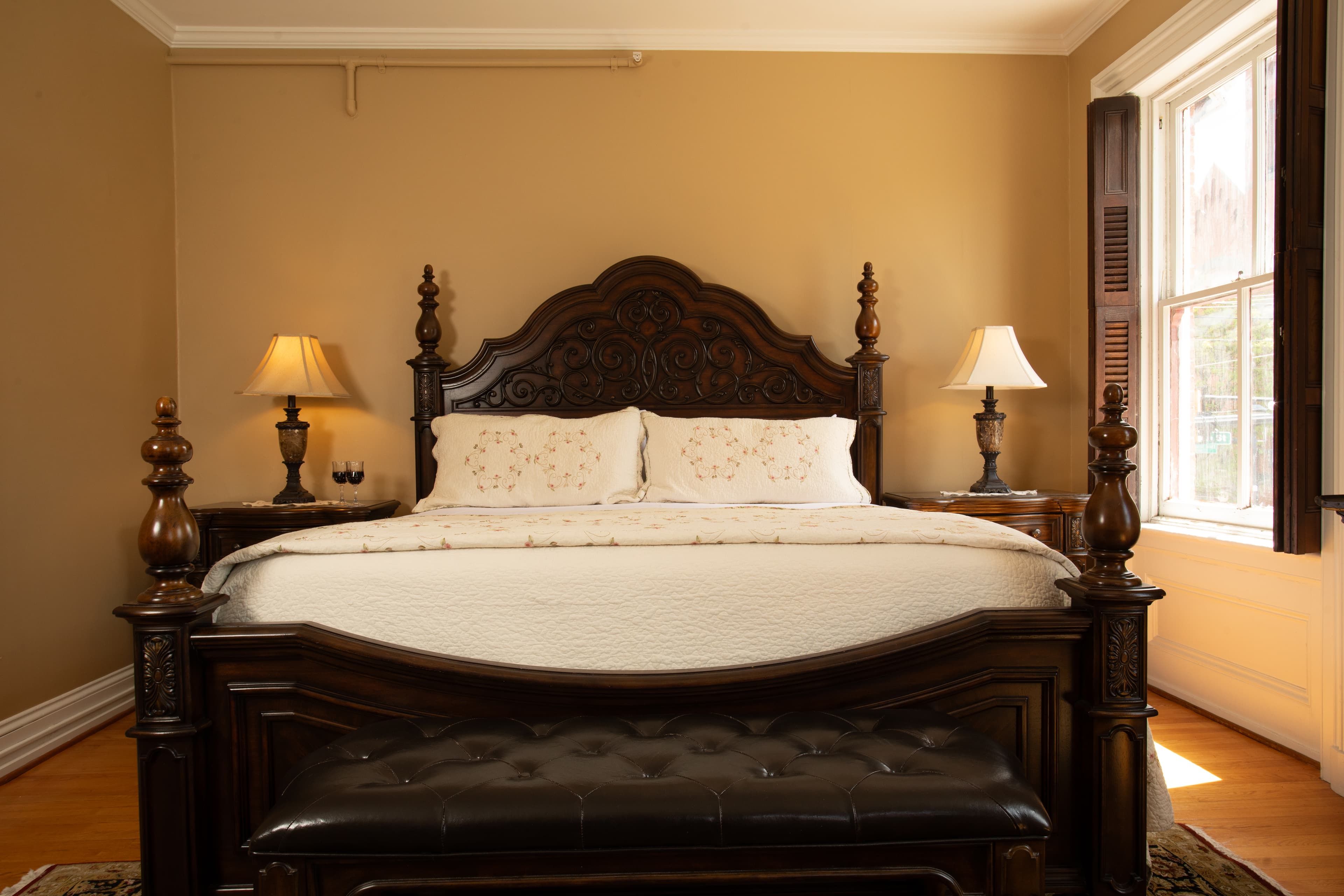 A beautifully styled bedroom with a large ornate wooden bed, bedside lamps, and a window letting in natural light.