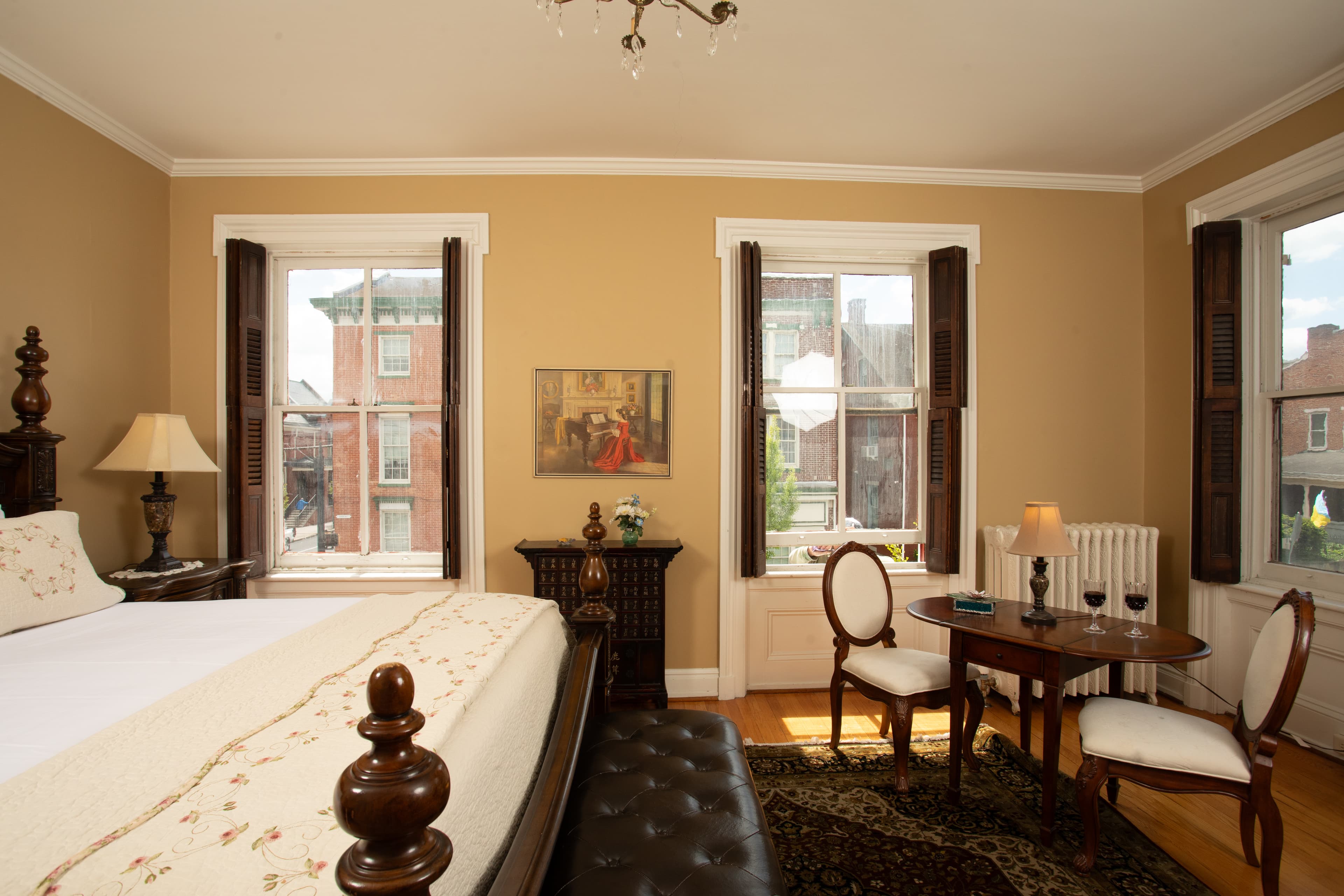 A cozy bedroom featuring a large bed, a small dining table, and two windows with light curtains.