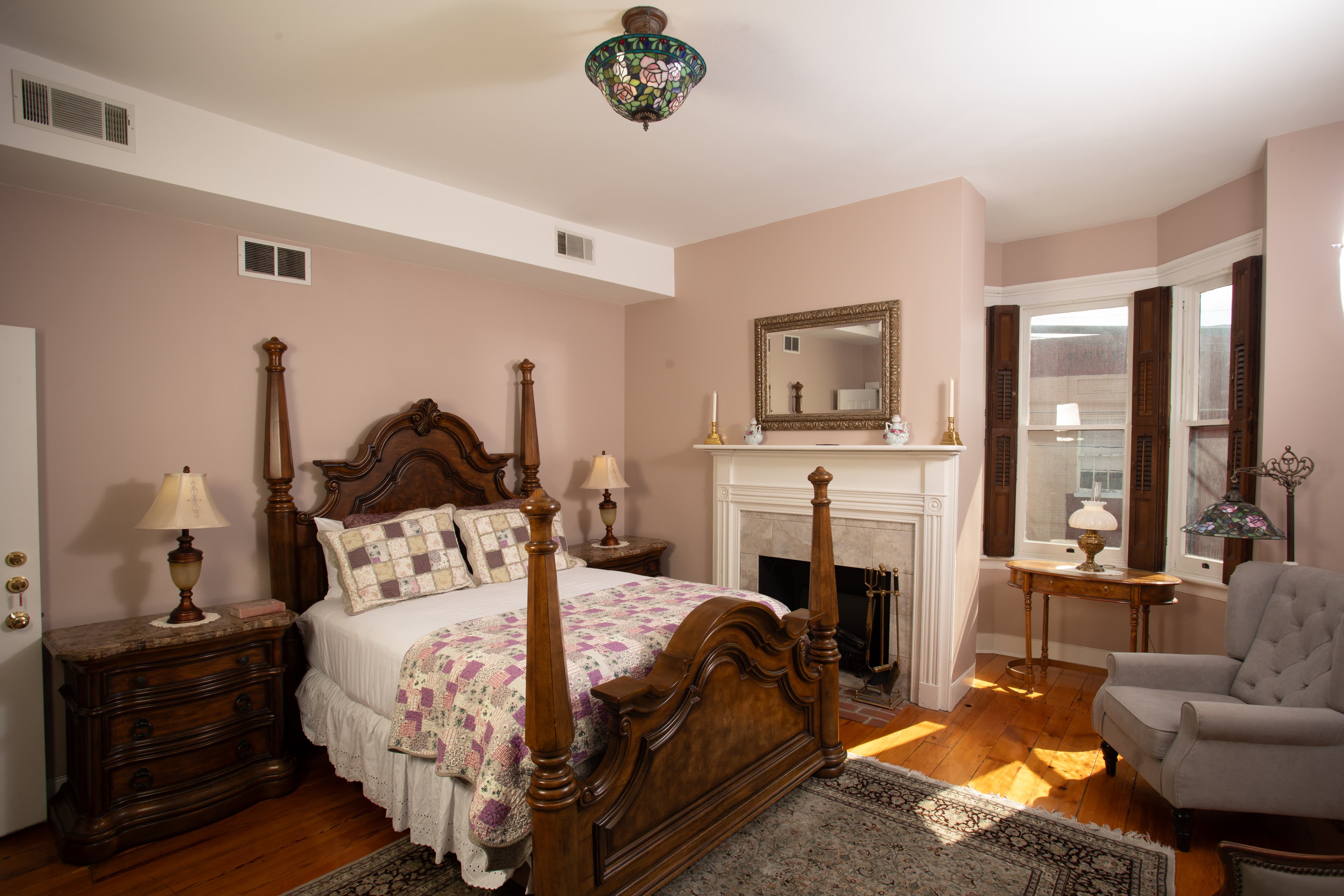 A cozy bedroom featuring a wooden bed, stylish lamps, a fireplace, and large windows.