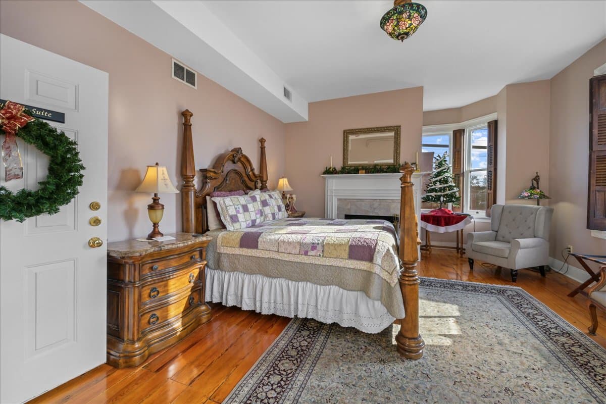 The Gettysburg Suite features an ornate wooden queen size poster bed, with 2 night stands with lamps, original wooden floors, and area rug. This room also features an electric fireplace, a leather settee, an armchair, a dresser, and TV.