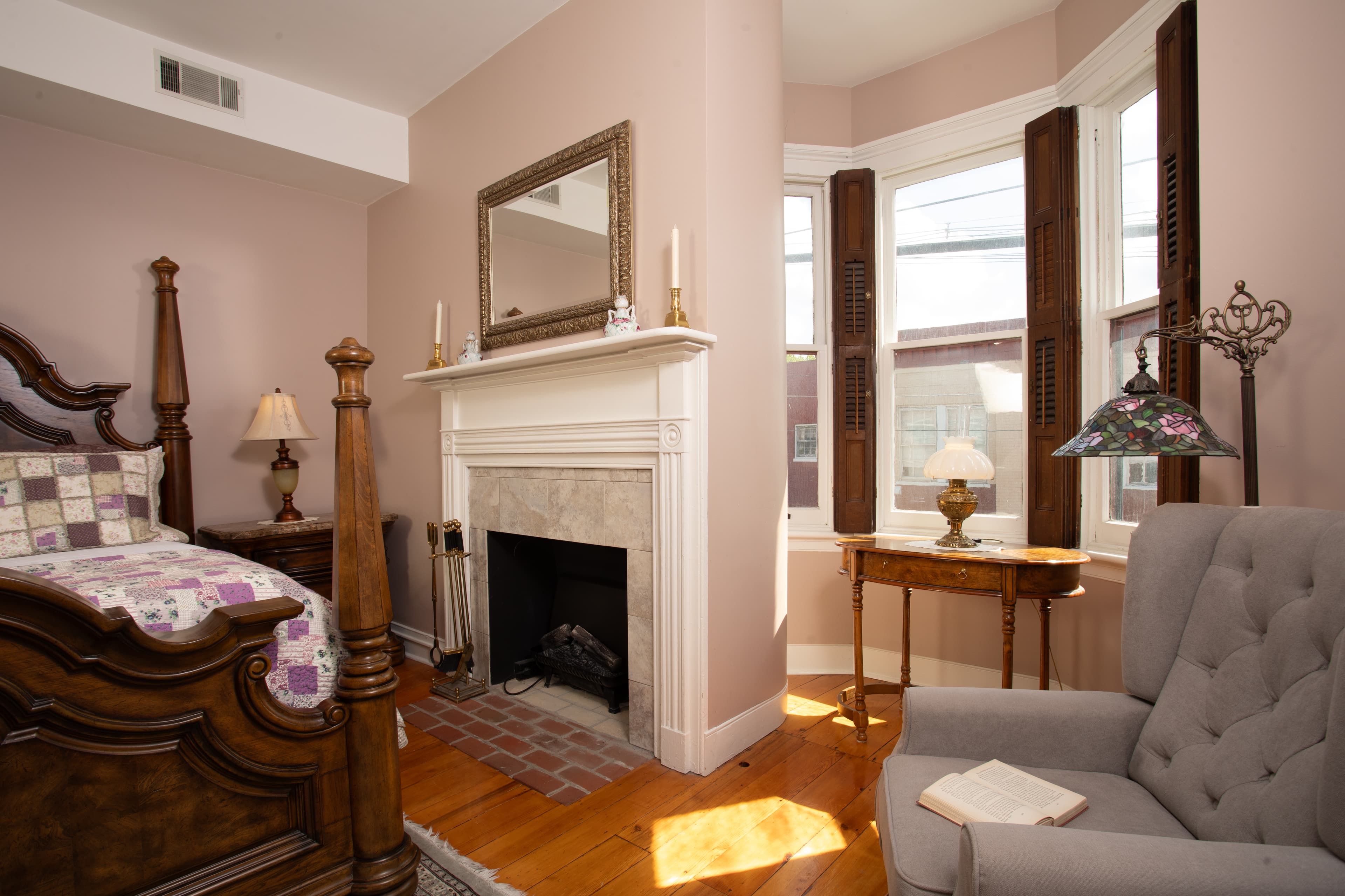 A cozy bedroom featuring a wooden bed, a fireplace with a mirror above it, and a comfortable armchair by a window.