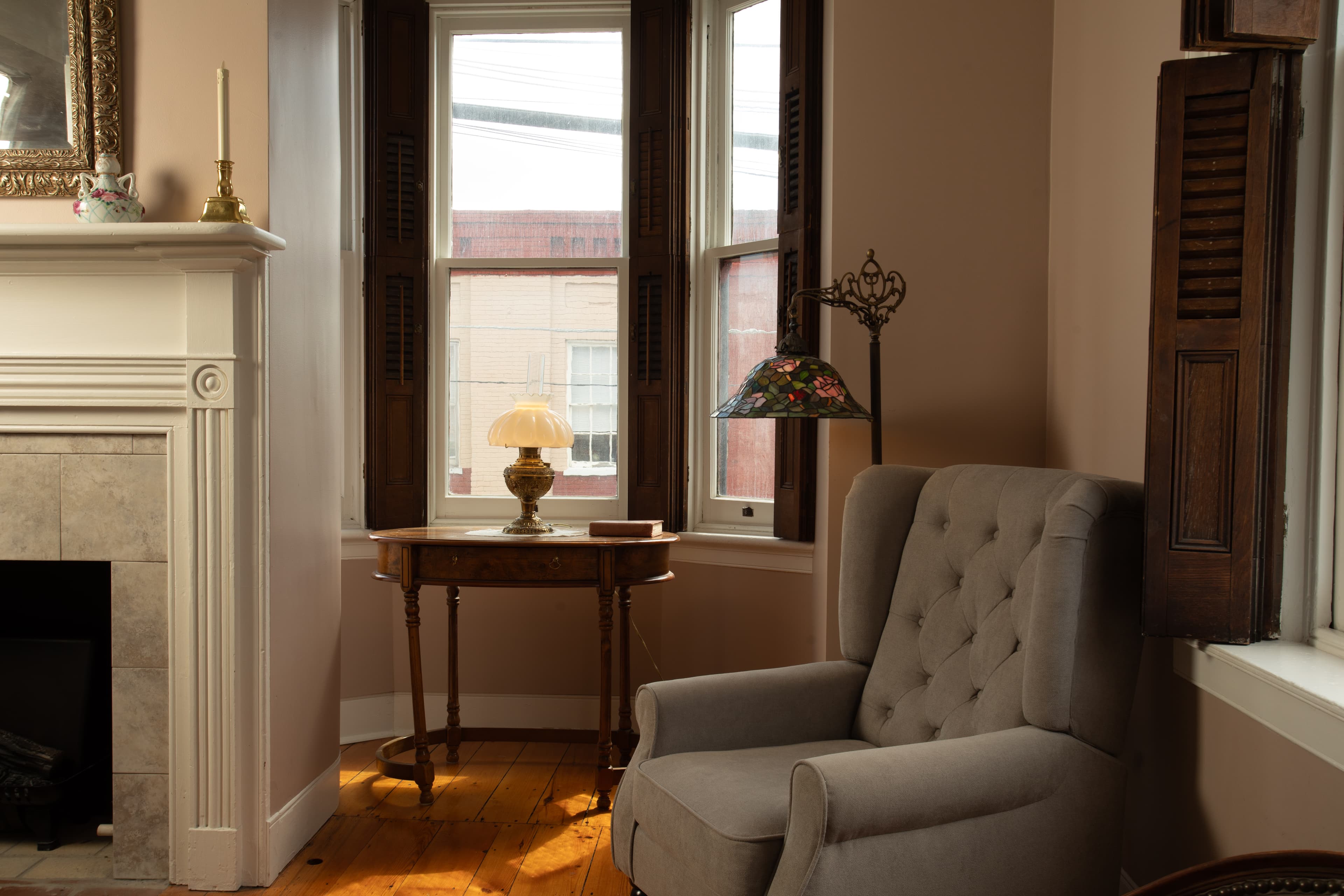 A light-colored armchair next to a bay window with striped curtains, a small table with a lamp, and wooden floors.