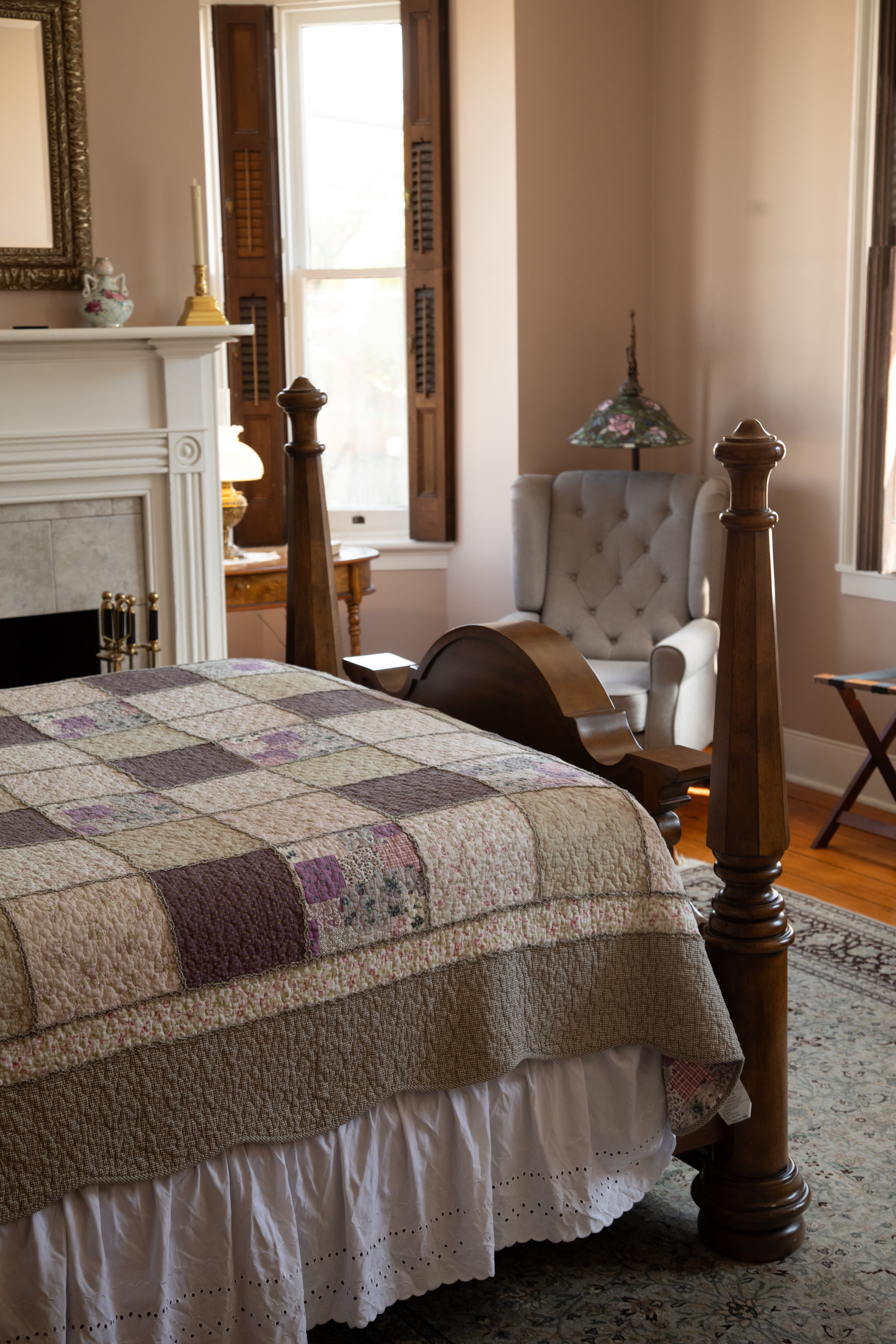 A cozy bedroom featuring a wooden four-poster bed with a patchwork quilt and a comfortable armchair beside a fireplace.
