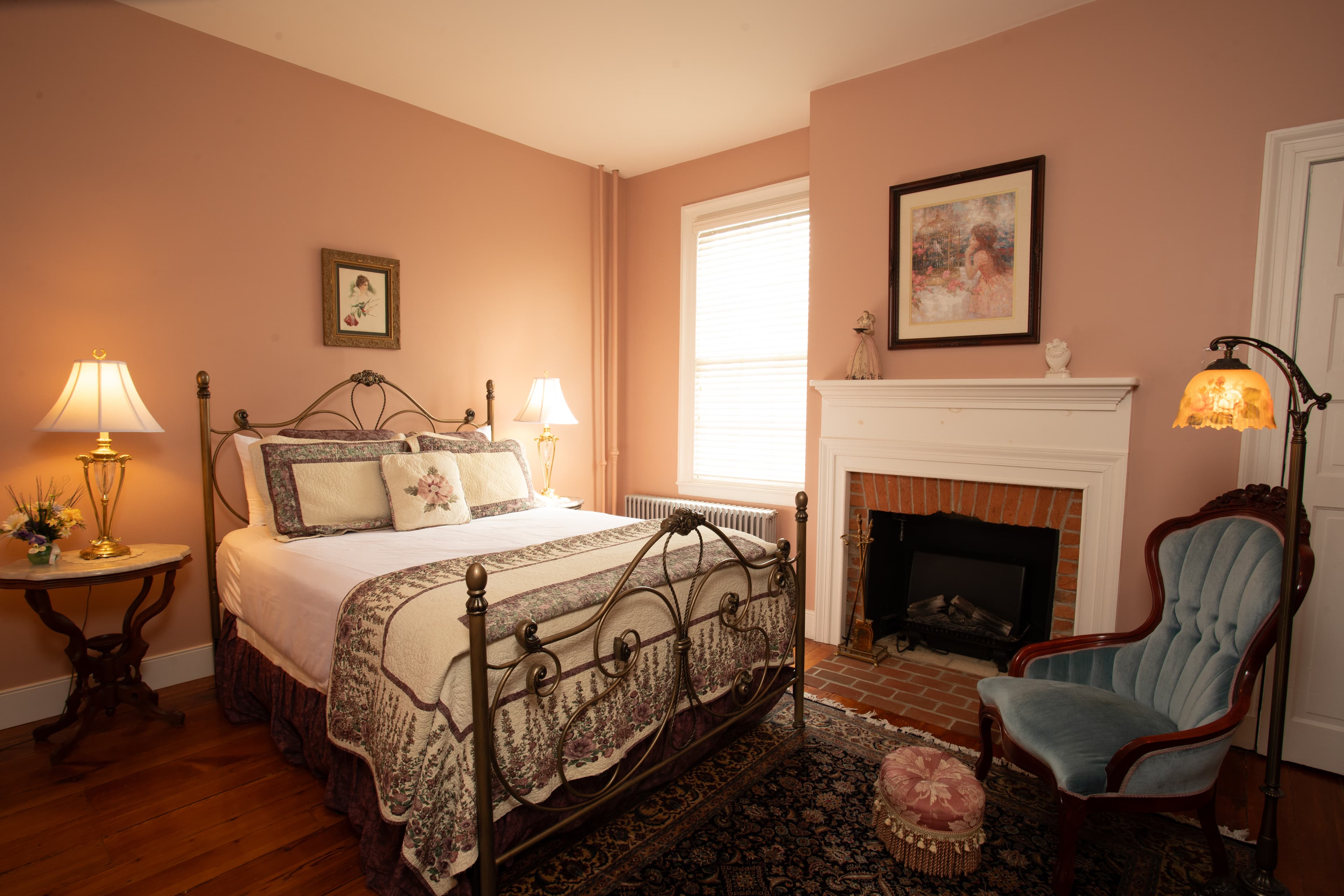 A decorated bedroom featuring a metal bed frame, bedside lamps, and a cozy armchair beside a fireplace.