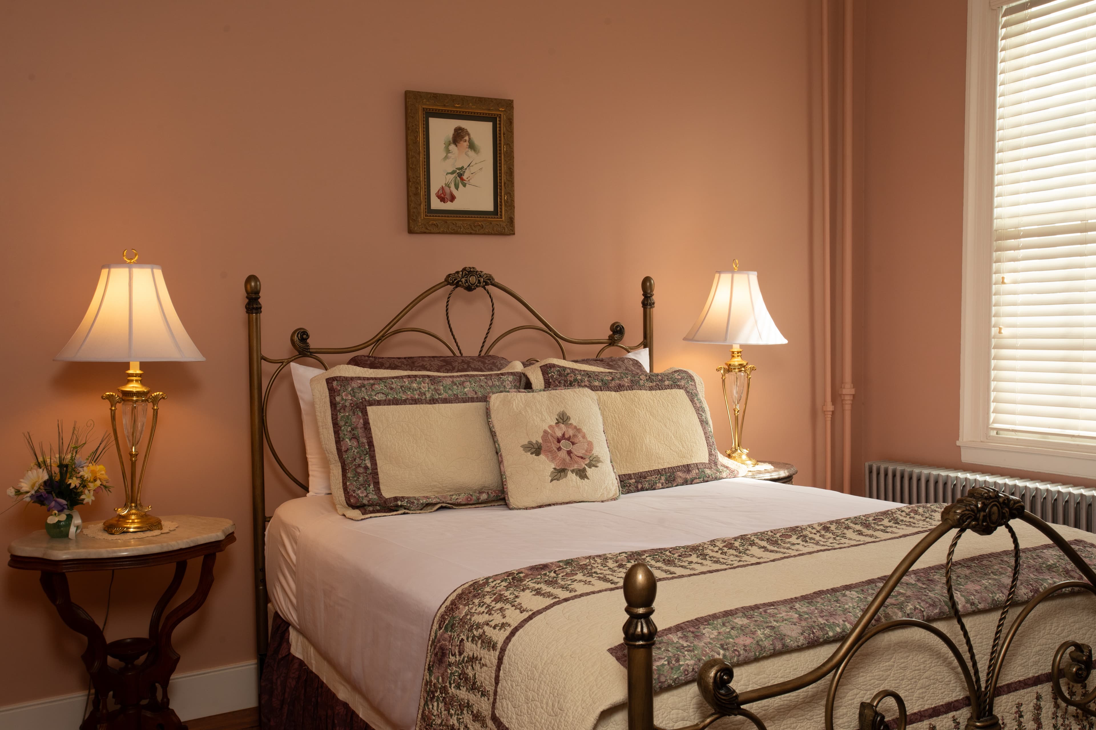 A cozy bedroom featuring a wrought iron bed, decorative pillows, and two bedside lamps against a soft pink wall.