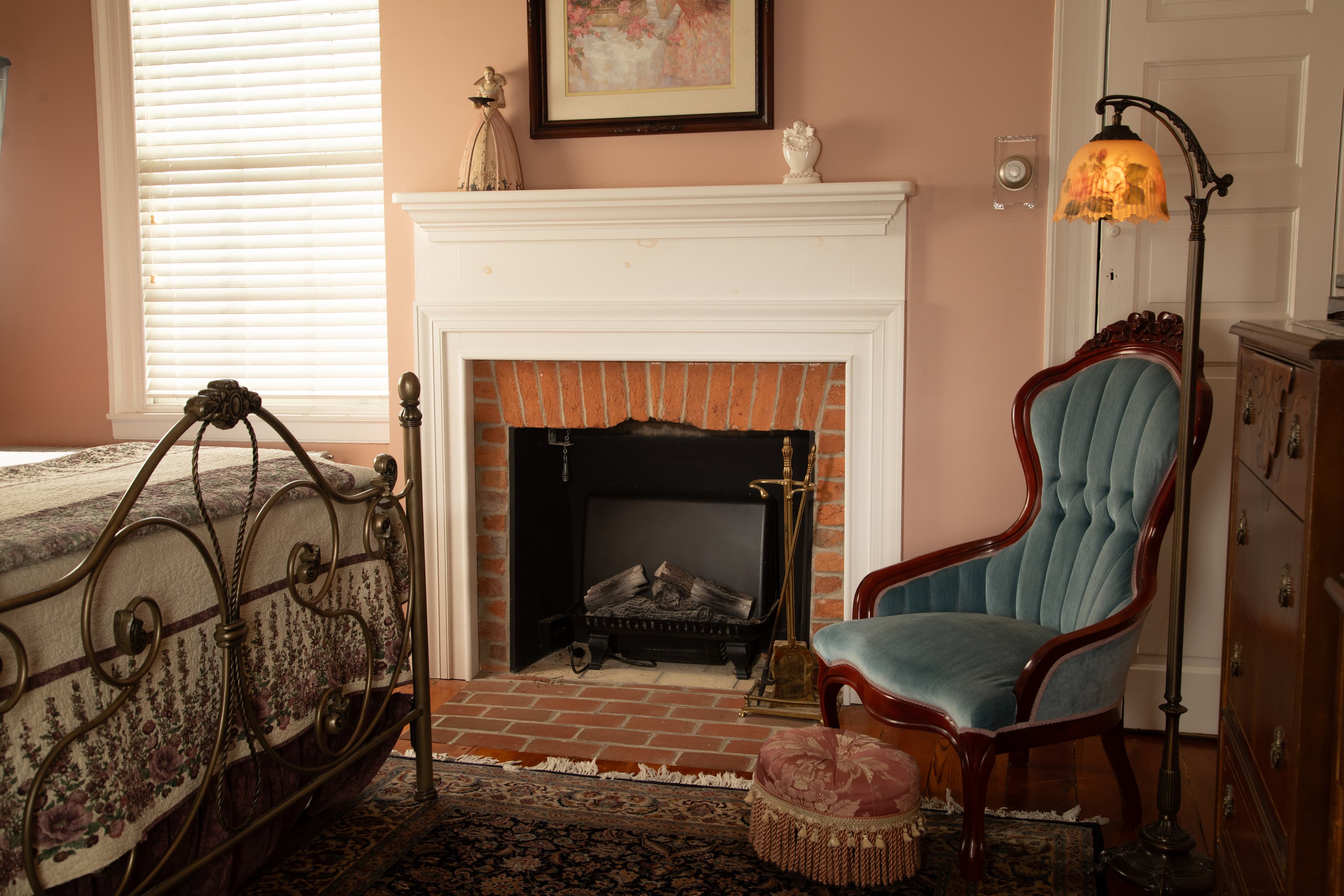 A cozy bedroom featuring a vintage iron bed, a blue upholstered chair, and a brick fireplace.