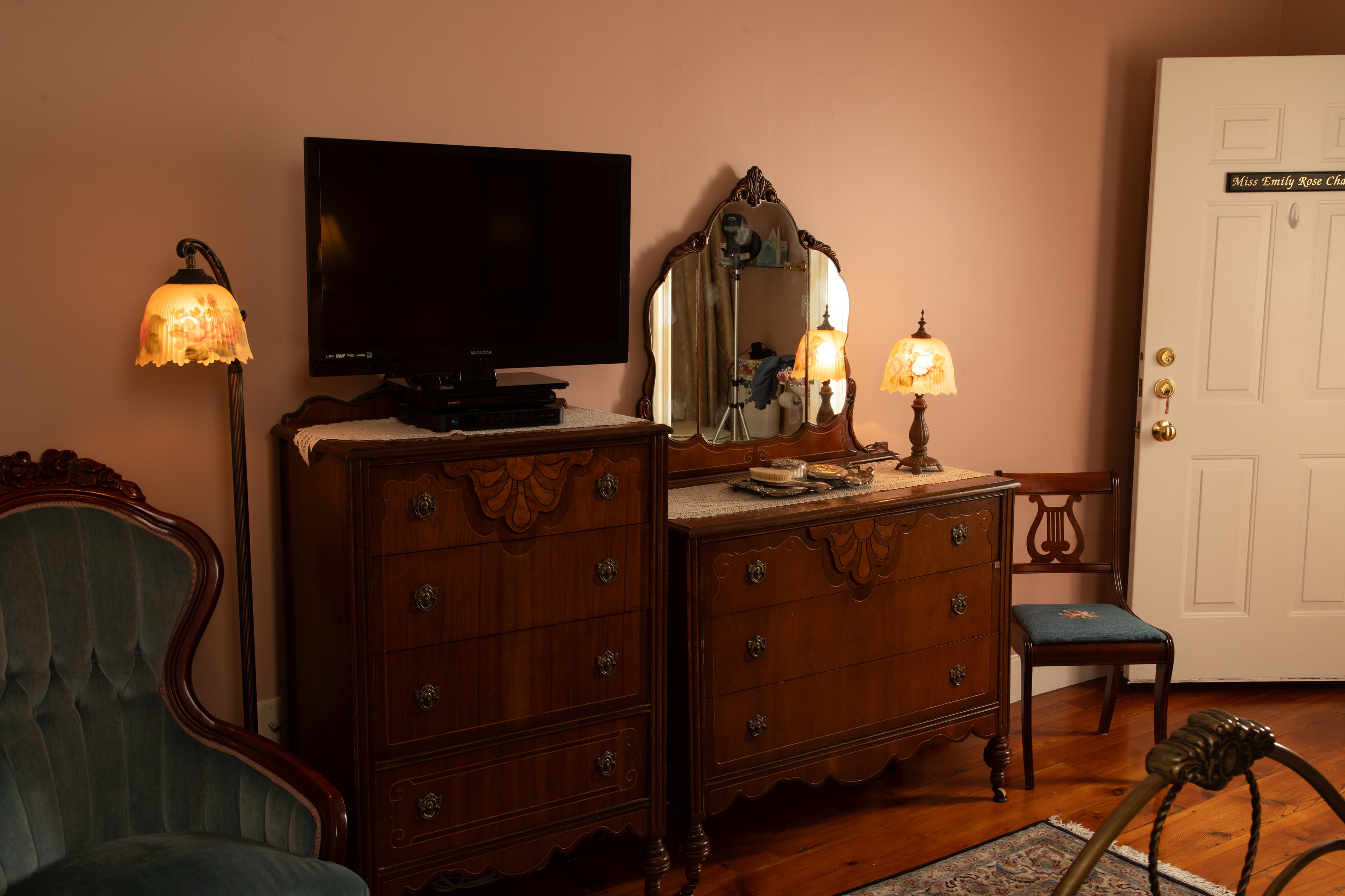 A cozy room featuring antique dressers, decorative lamps, a wall-mounted TV, and a door leading outside.