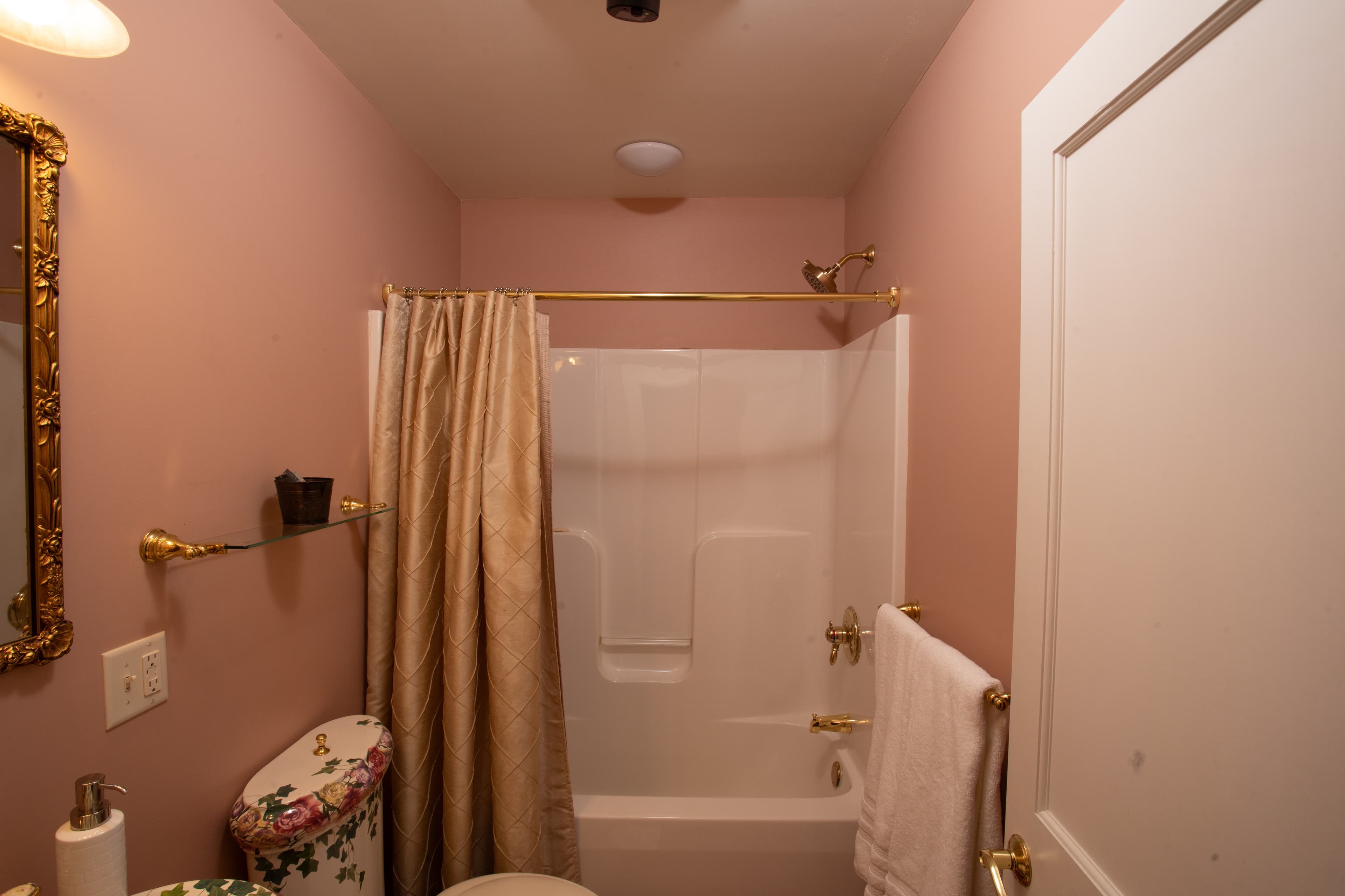 A bathroom featuring a pink wall, shower with curtain, decorative towel, and a floral toilet cover.