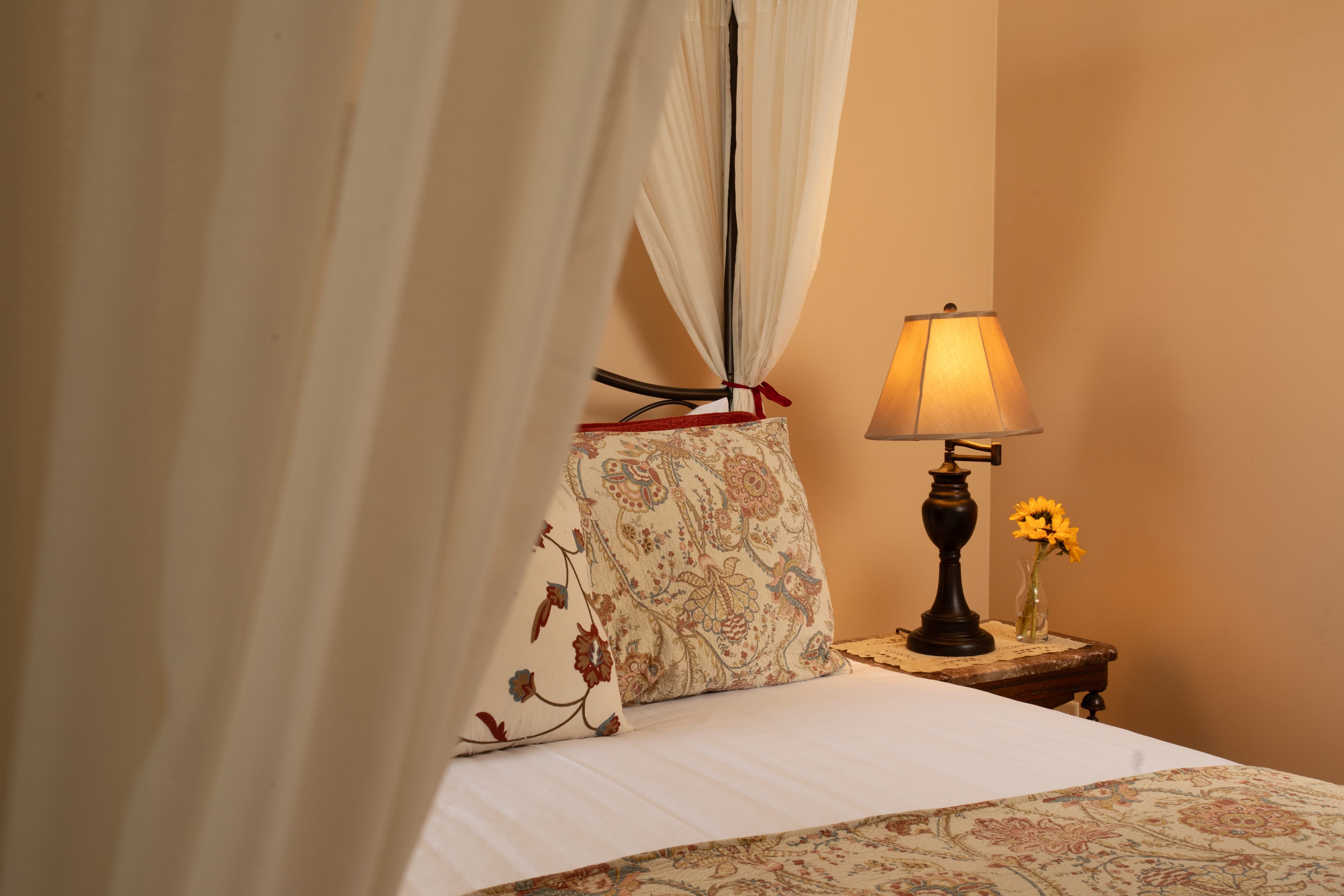 A cozy bedroom featuring a bed with decorative pillows, a bedside lamp, and a small vase with a sunflower.
