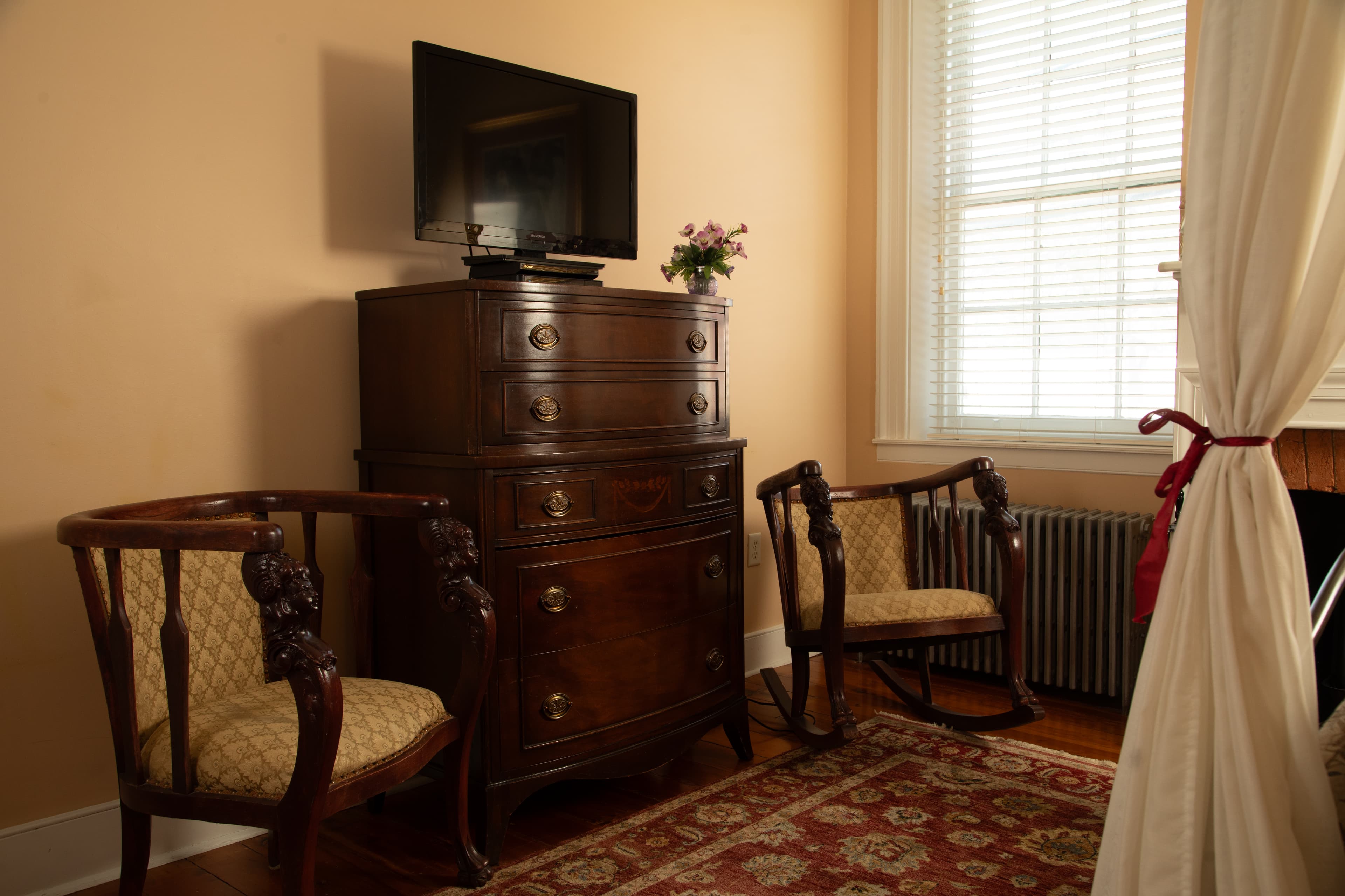 A cozy room featuring a wooden dresser with a TV on top, two ornate chairs, and a window with light drapes.