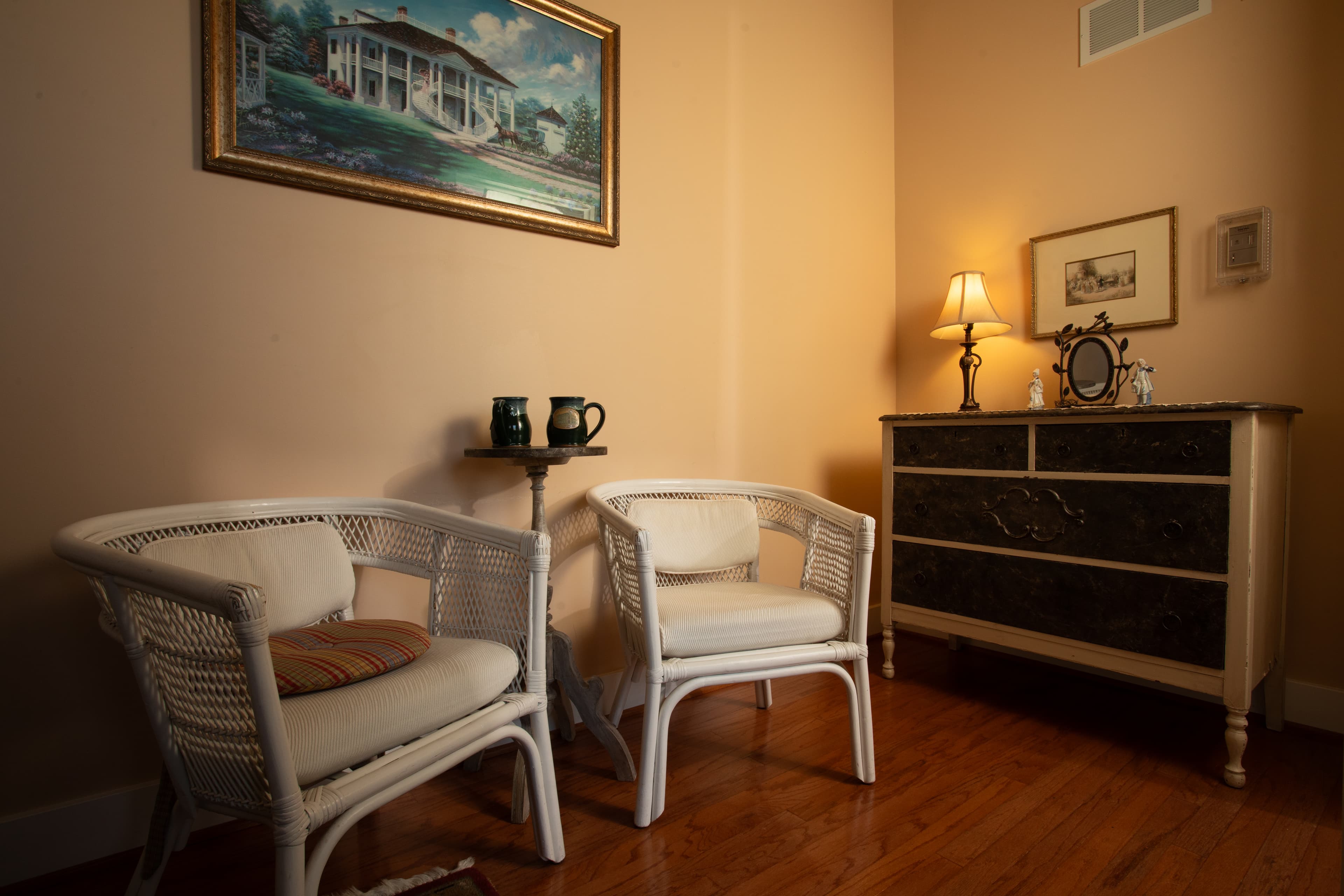 A cozy nook featuring two white wicker chairs, a small table with mugs, a dresser, and a framed painting on the wall.