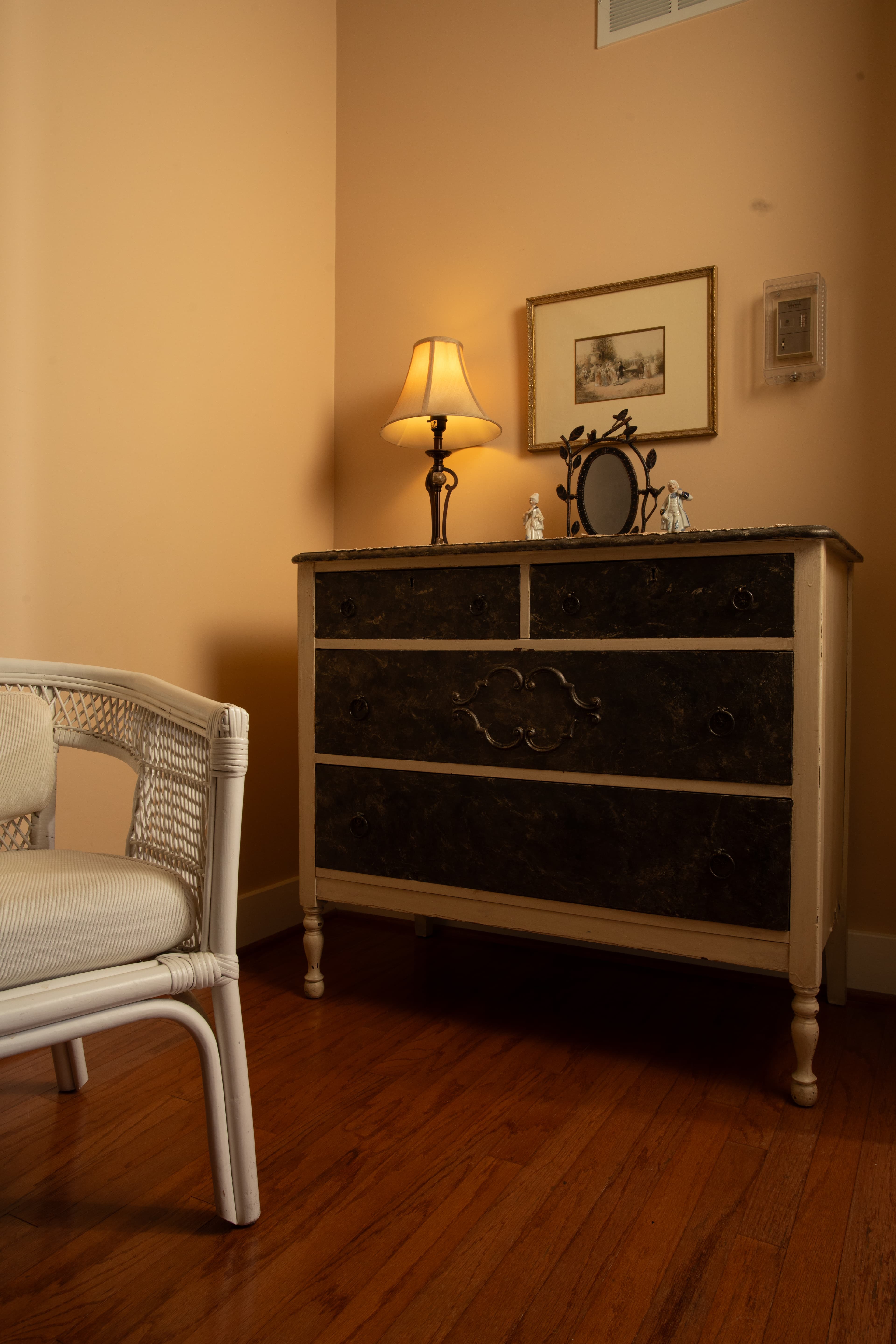 A vintage dresser with a lamp and a mirror sits in a warmly lit corner of a room next to a wicker chair.