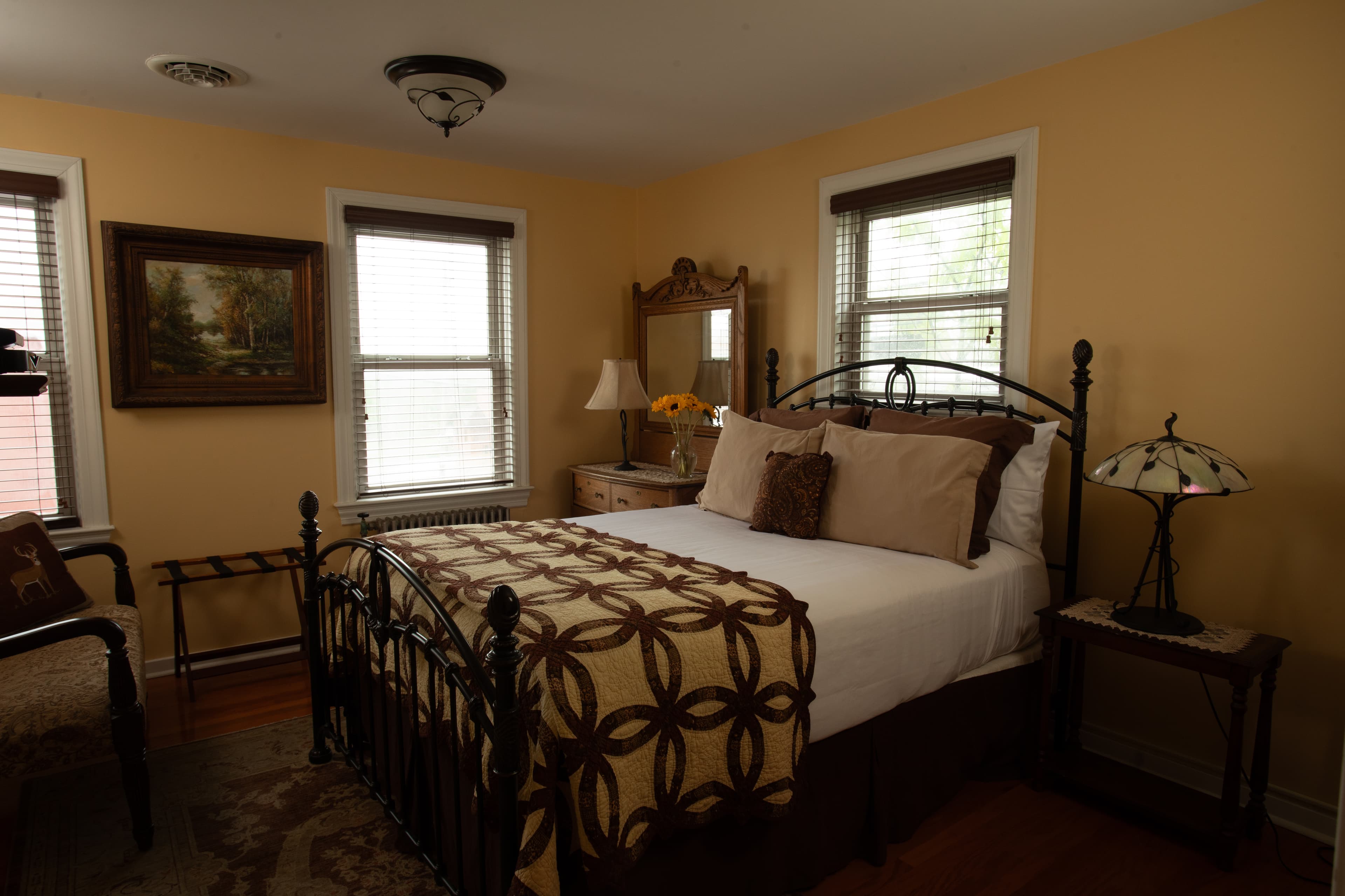 A cozy, well-furnished bedroom with a metal frame bed, warm yellow walls, and natural light from three windows.