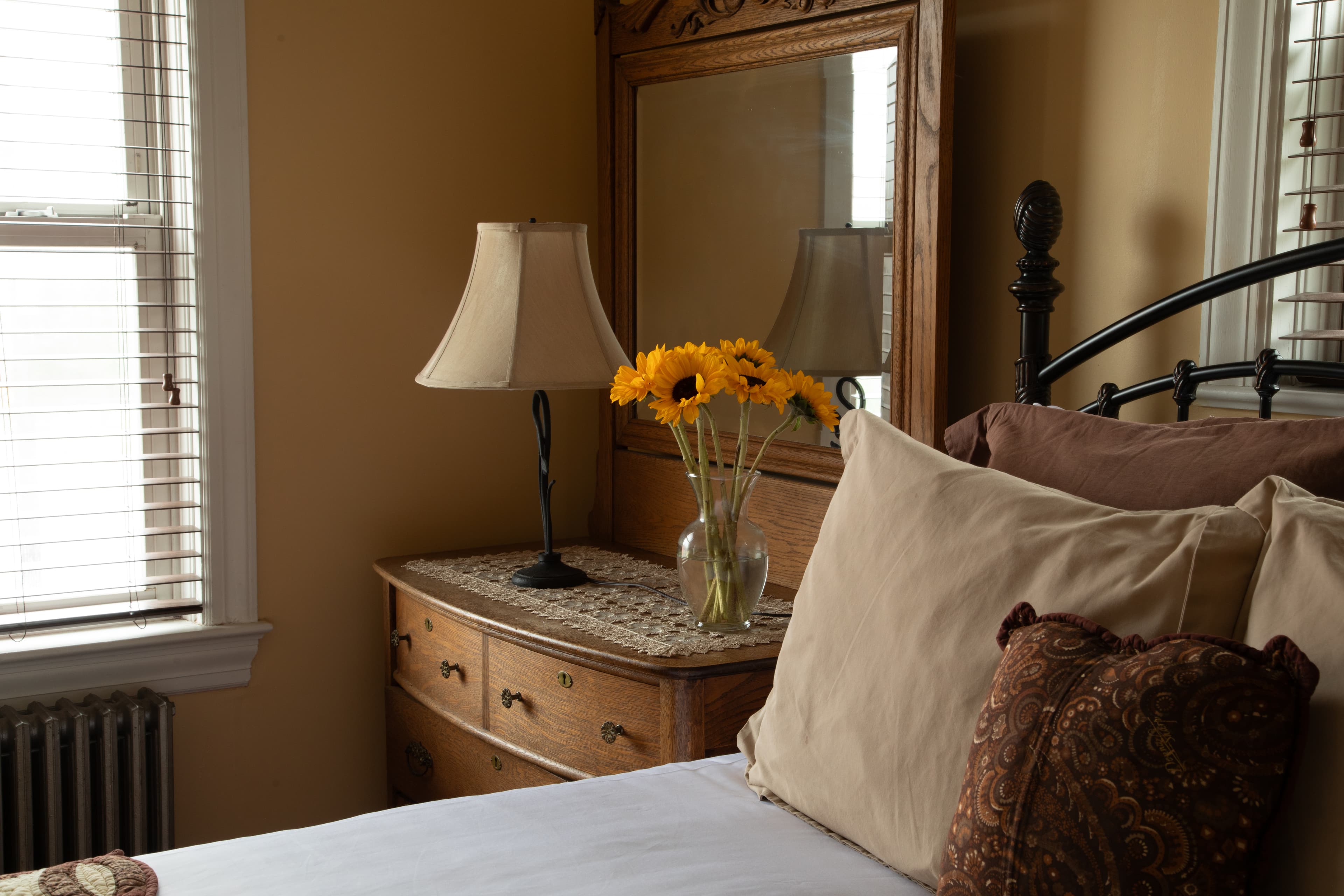 A cozy bedroom featuring a wooden nightstand with a lamp and a vase of yellow flowers beside a neatly made bed.