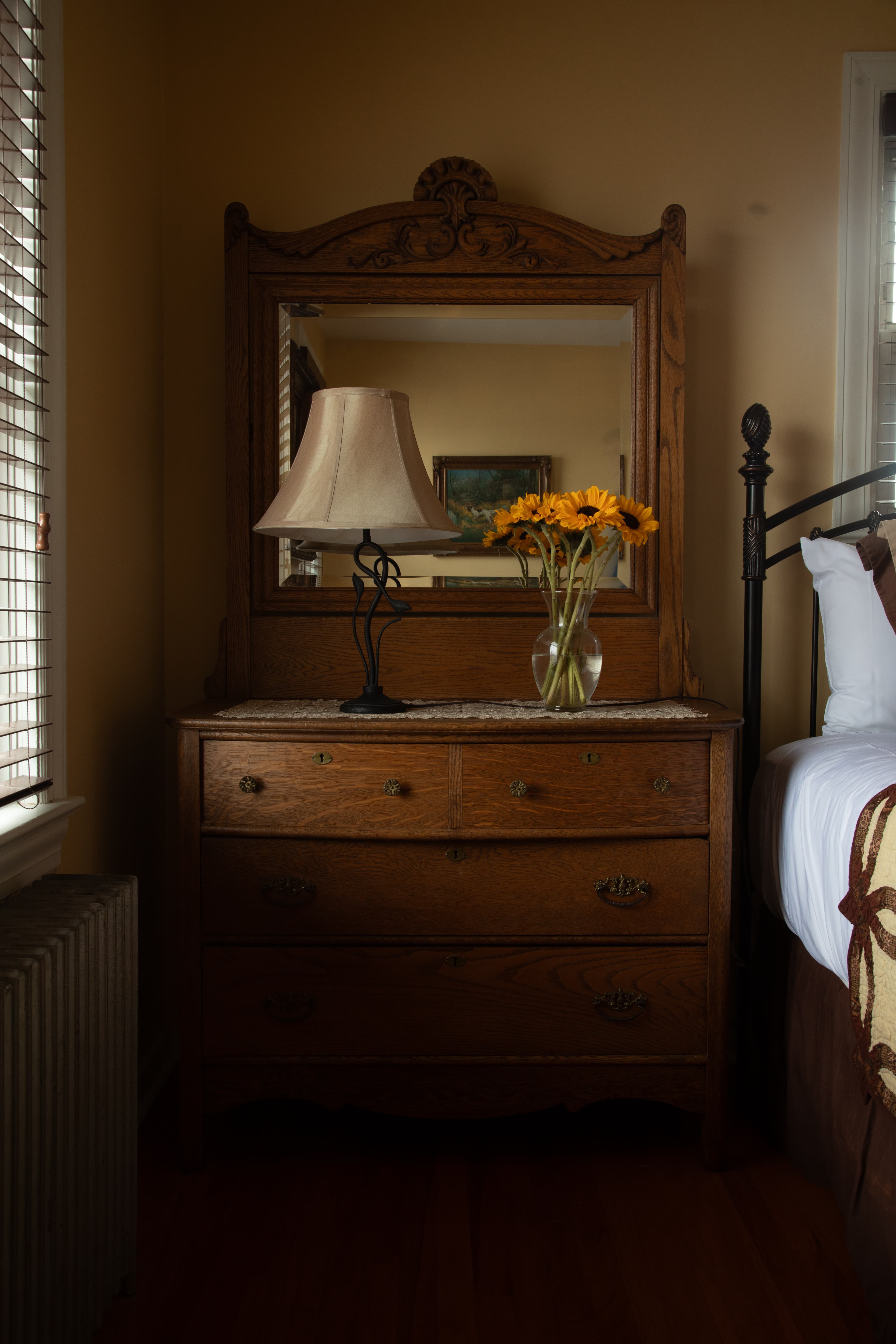A vintage wooden dresser with a mirror, a lamp, and a vase of yellow flowers in a warmly lit bedroom.