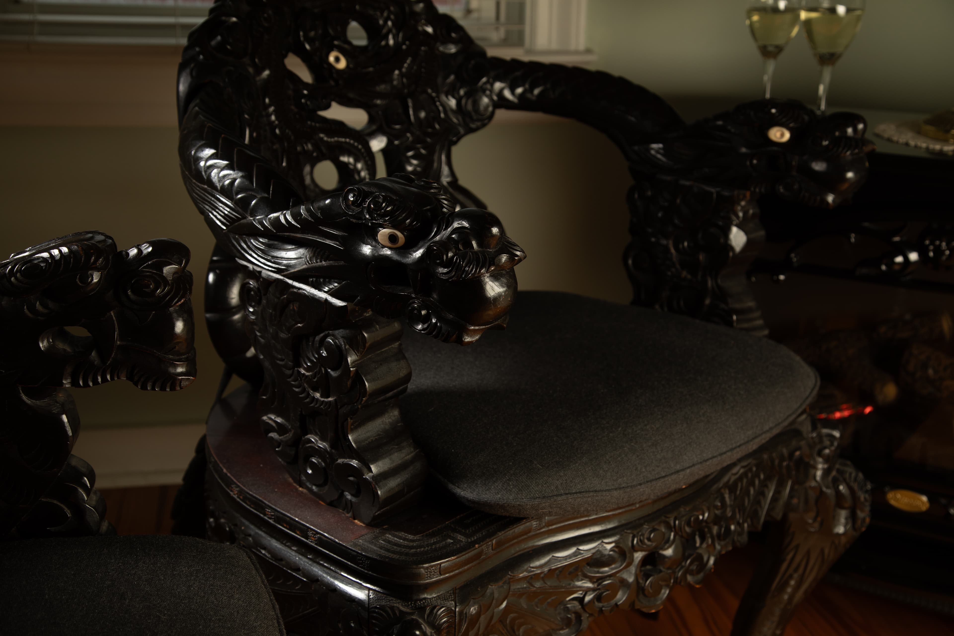 Intricately carved black wooden chair with grey cushion, near two glasses of wine.