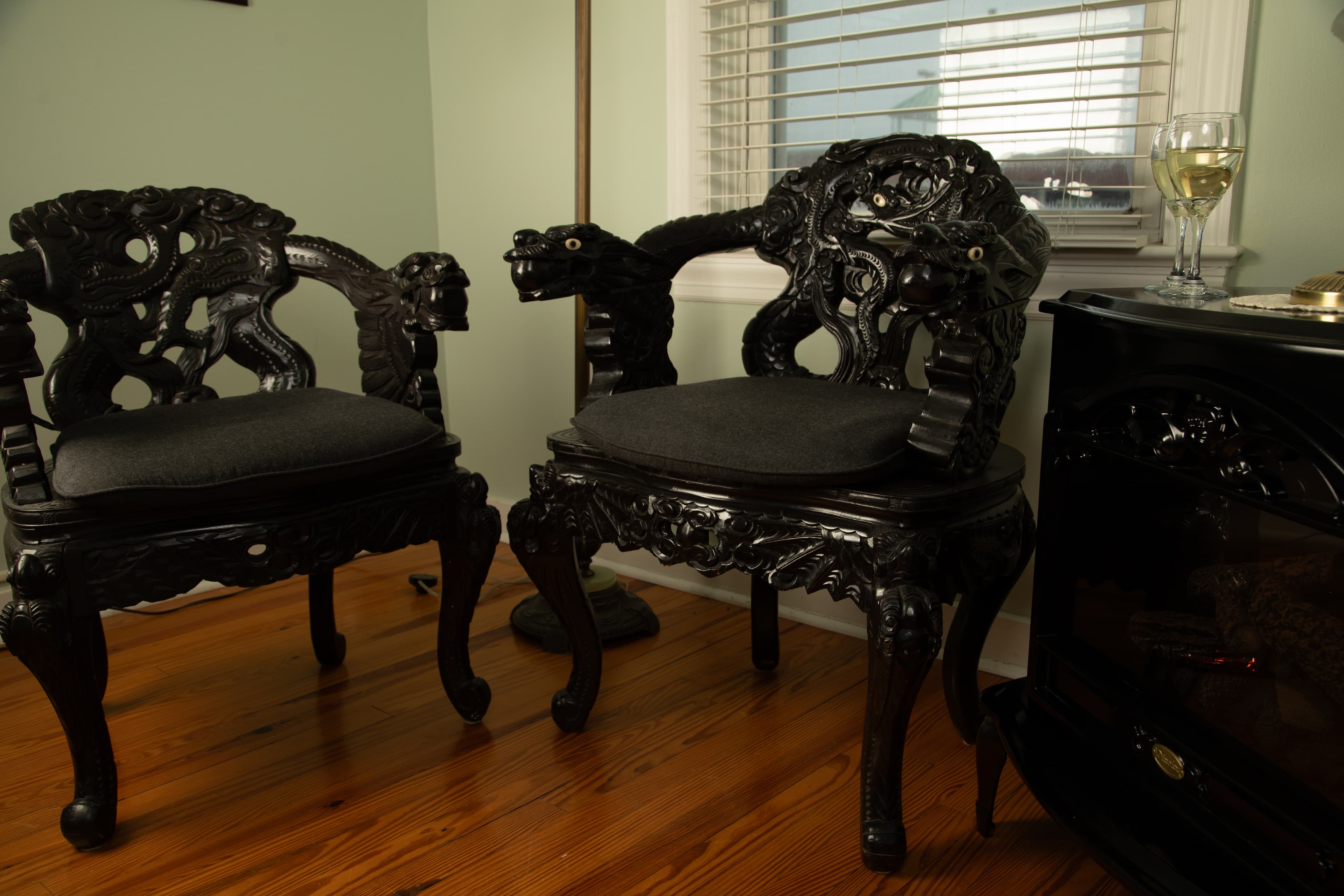 Two intricately carved black wooden chairs with dark cushions are positioned near a window and a glass of wine in the Cabernet Chamber.