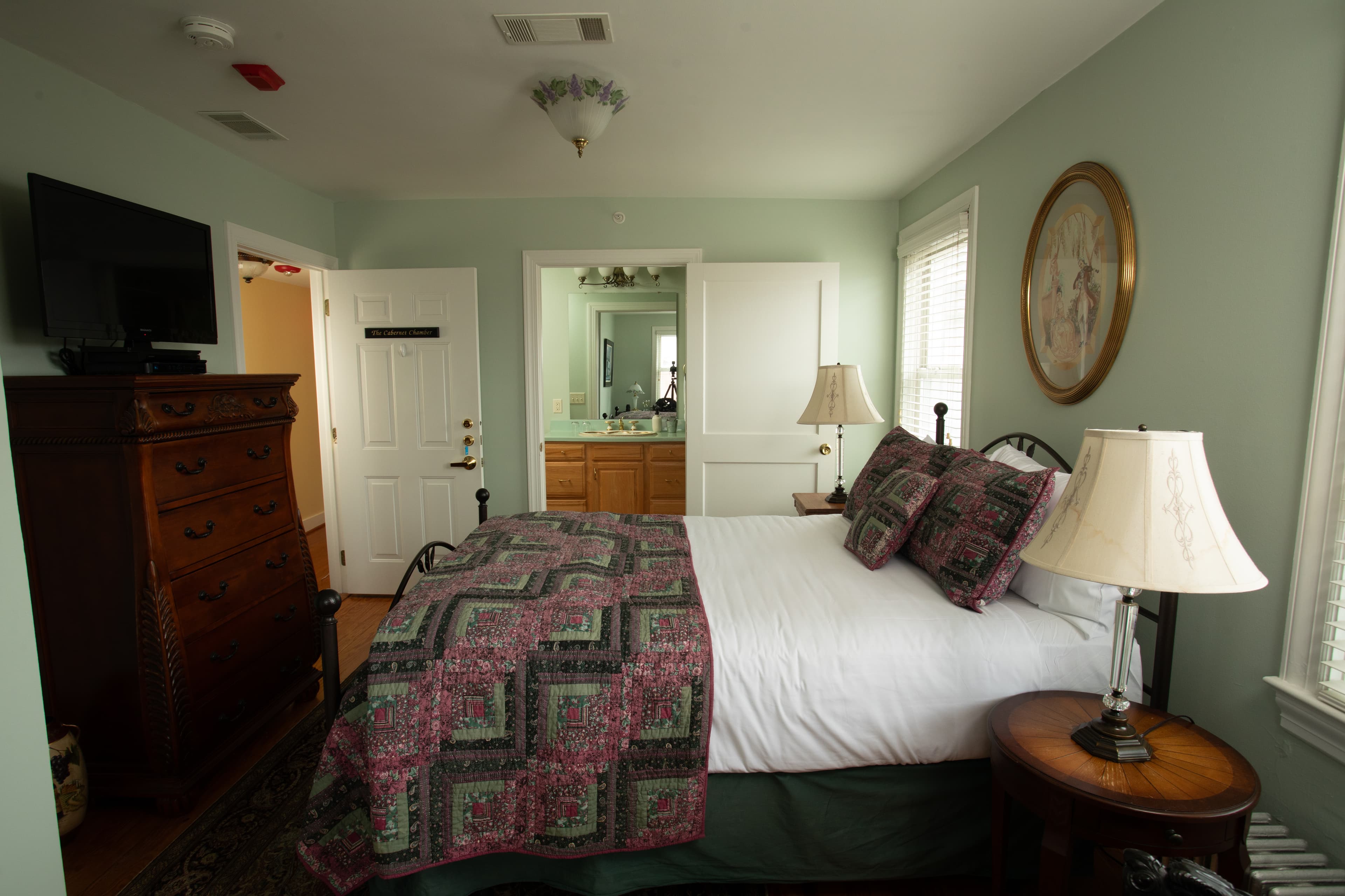 A cozy bedroom, Cabernet Chamber, featuring a double bed with a patterned quilt, a wooden dresser, and a lamp, set against green walls.
