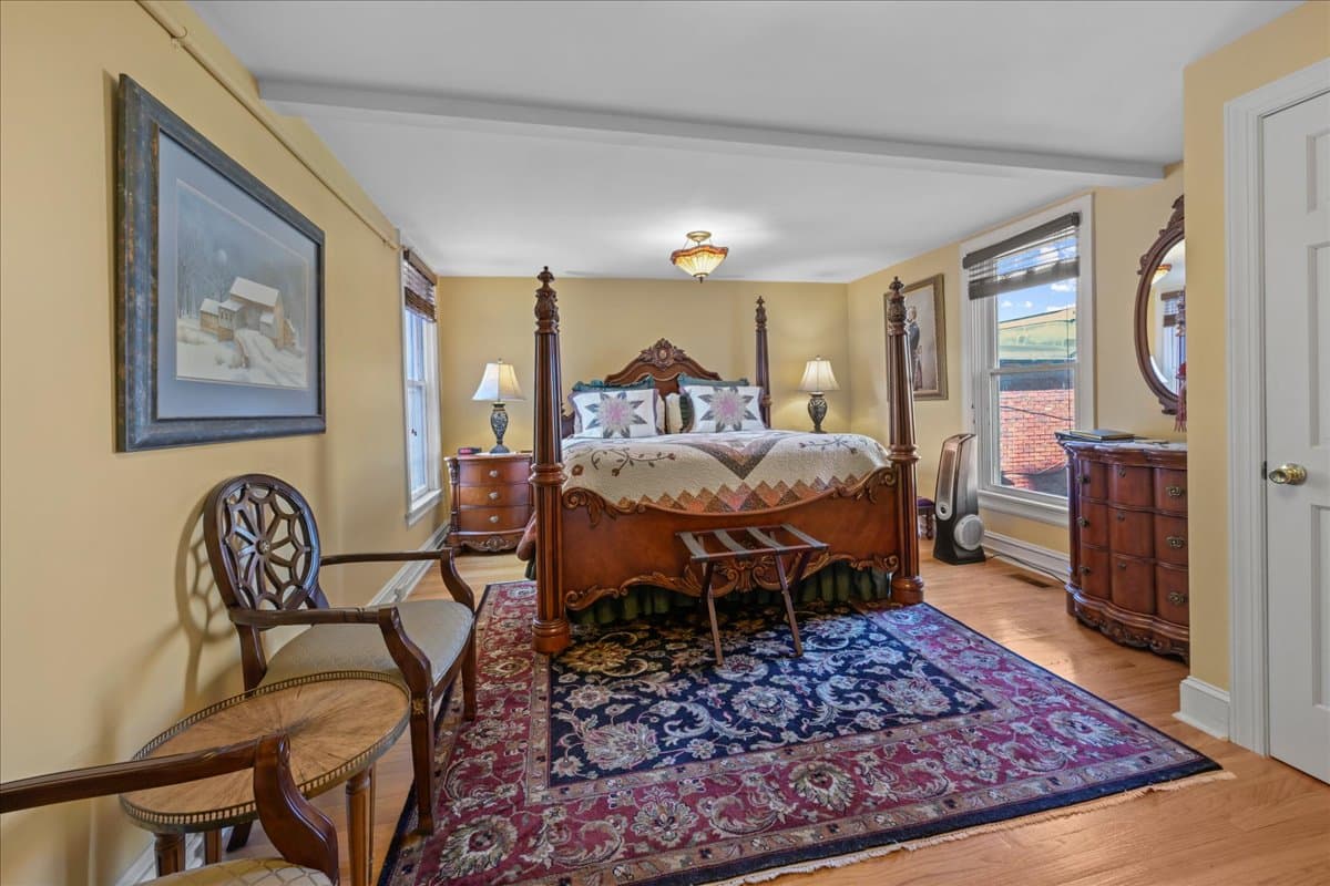 The Colonel Chamberlain Suite features a large ornate king-size wooden poster bed, 2 night stands with lamps, 2 chairs, and large wooden electric fireplace.