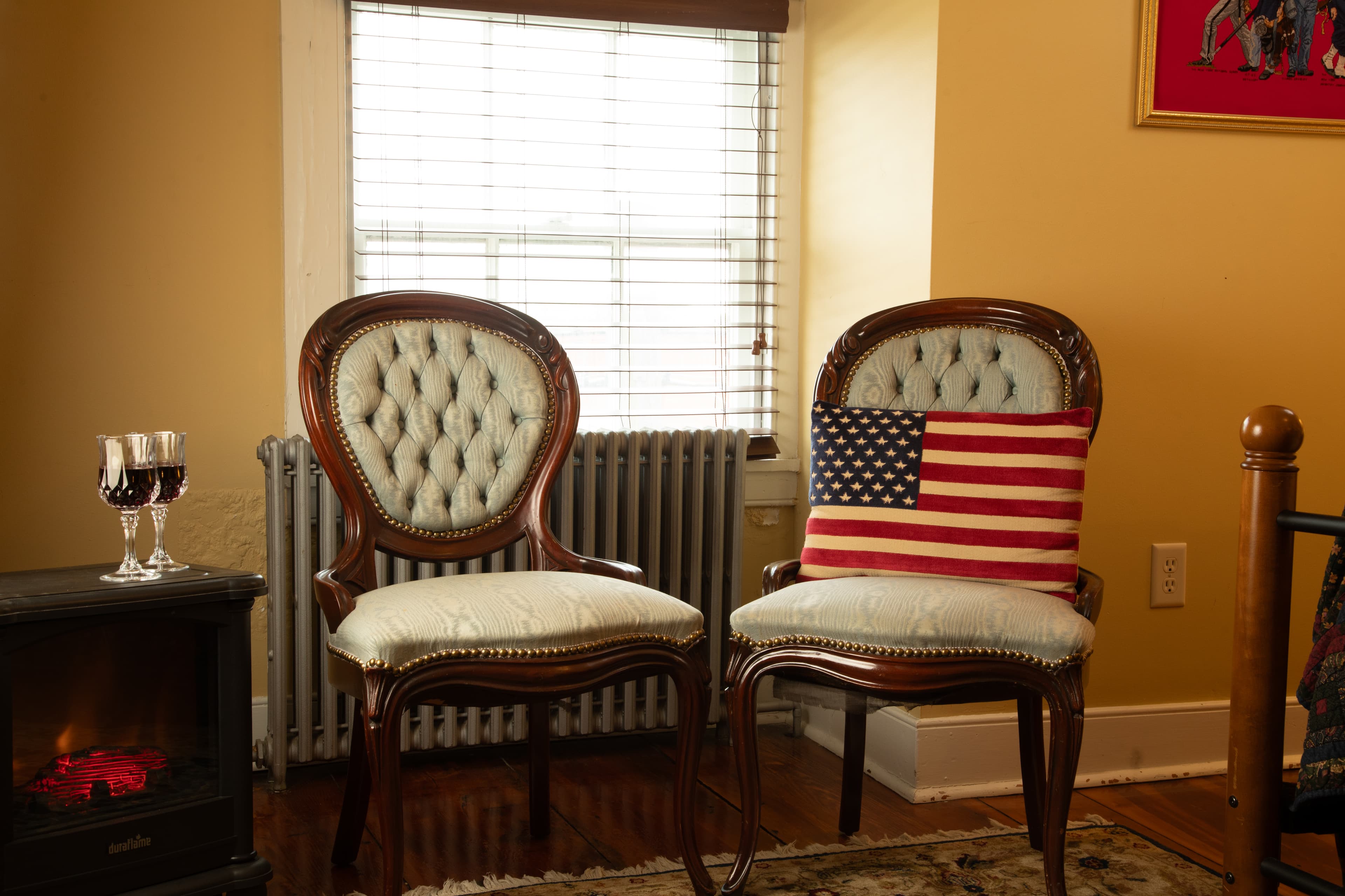 A cozy corner features two elegant chairs, one adorned with a pillow displaying the American flag, next to a small table with wine glasses and a decorative heater.