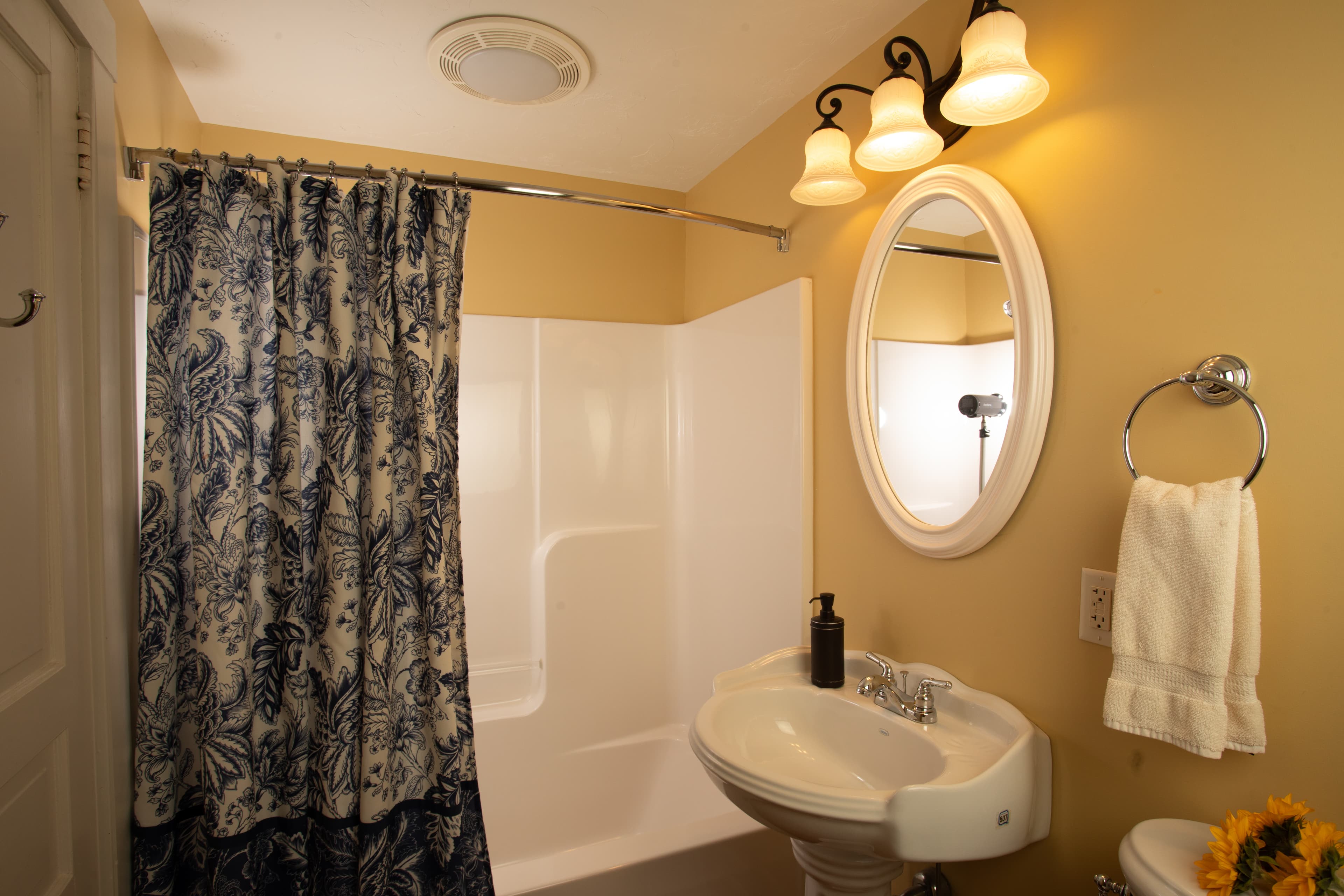 A small bathroom featuring a shower with a floral curtain, a sink, a round mirror, and decorative lighting.