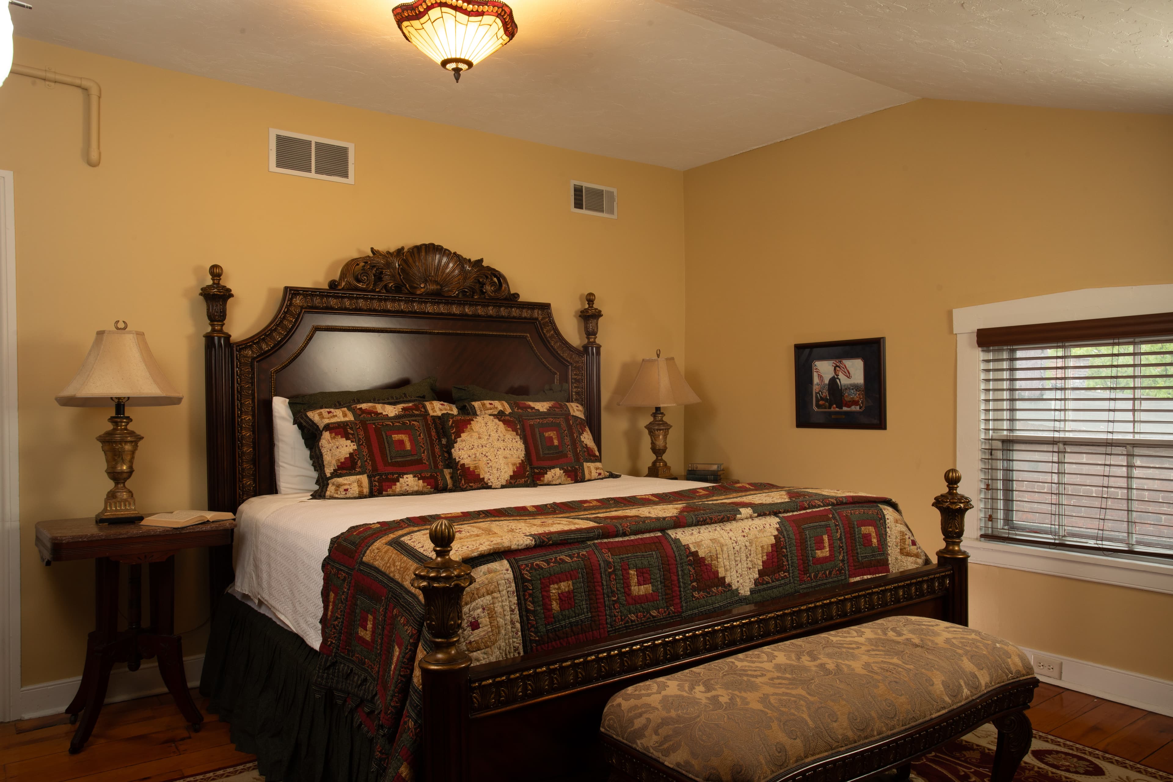 The Lincoln Suite features an Ornate wooden King size bed, 2 Victorian night stands with lamps, a bench at the foot of the bed, a dresser, a desk with chair, a small refrigerator, original wooden floors with an area rug.