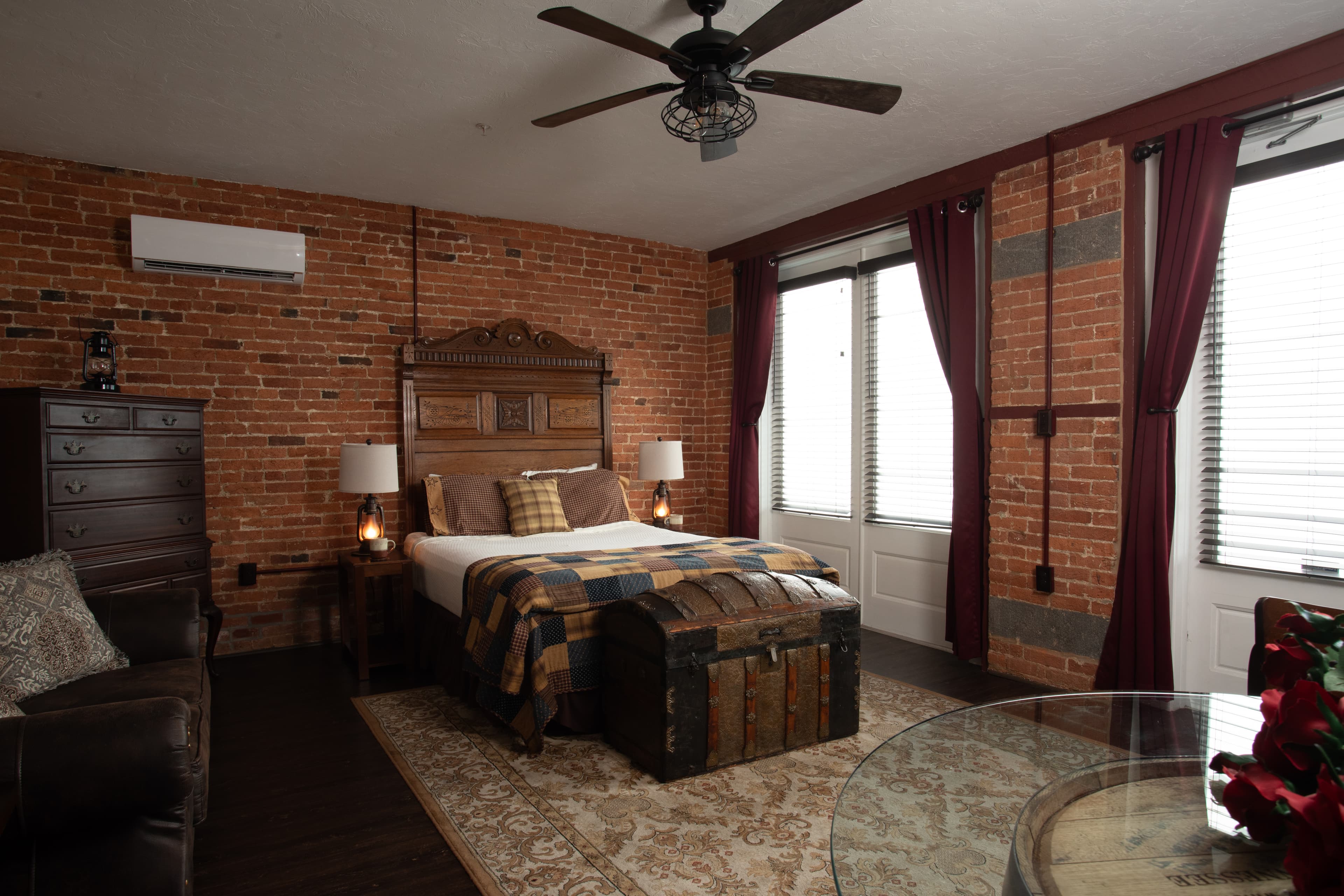 The Carriage House bedroom featuring exposed brick walls, a bed with plaid bedding, and large windows with red curtains.